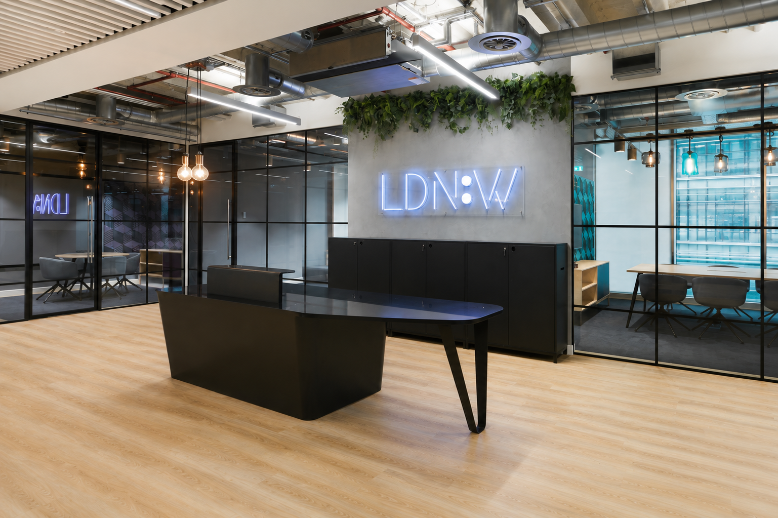 Modern office reception area with black desk, glass-walled meeting rooms, neon sign reading "LDNW," indoor plants, and contemporary lighting fixtures.
