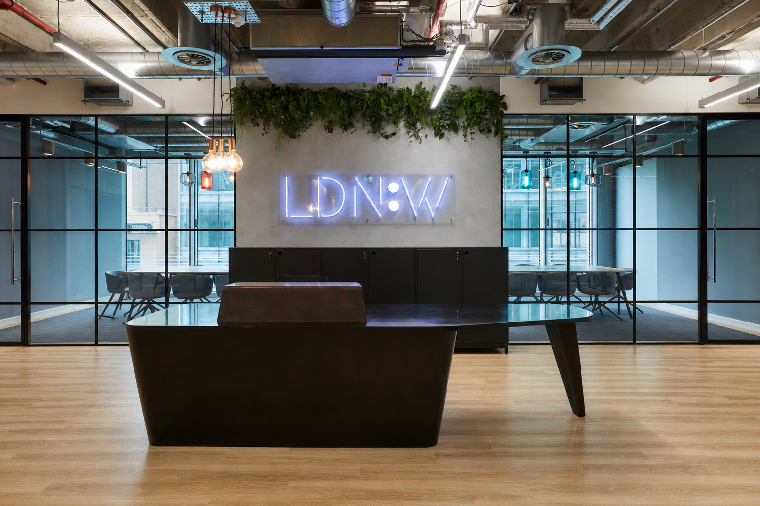 Modern office reception area with a black desk, a neon sign reading 'LDNW' on the wall, glass-walled meeting rooms in the background, and ceiling pipes and lighting fixtures.