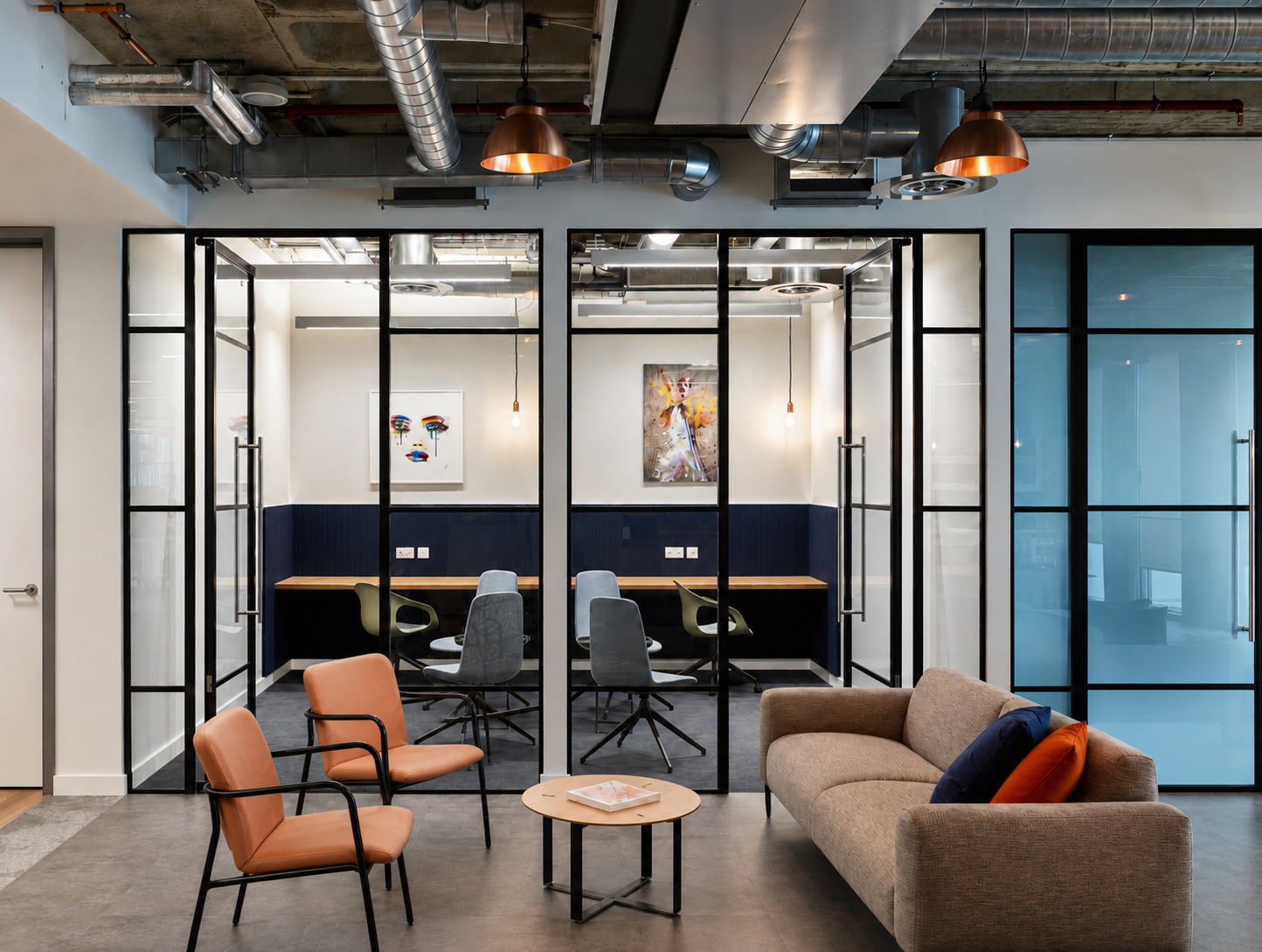 Modern office lounge area with two peach-colored chairs, a beige sofa with colorful pillows, and a small round table with a magazine on it. Behind the lounge, there is a glass-walled private meeting room with abstract art on the walls and office chai