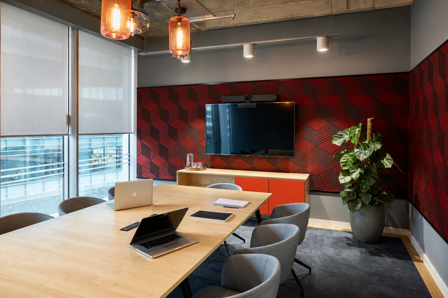 Modern conference room with a large wooden table, six gray chairs, a flat-screen TV mounted on a red and black geometric-patterned wall, a big potted plant, and large windows with white shades.