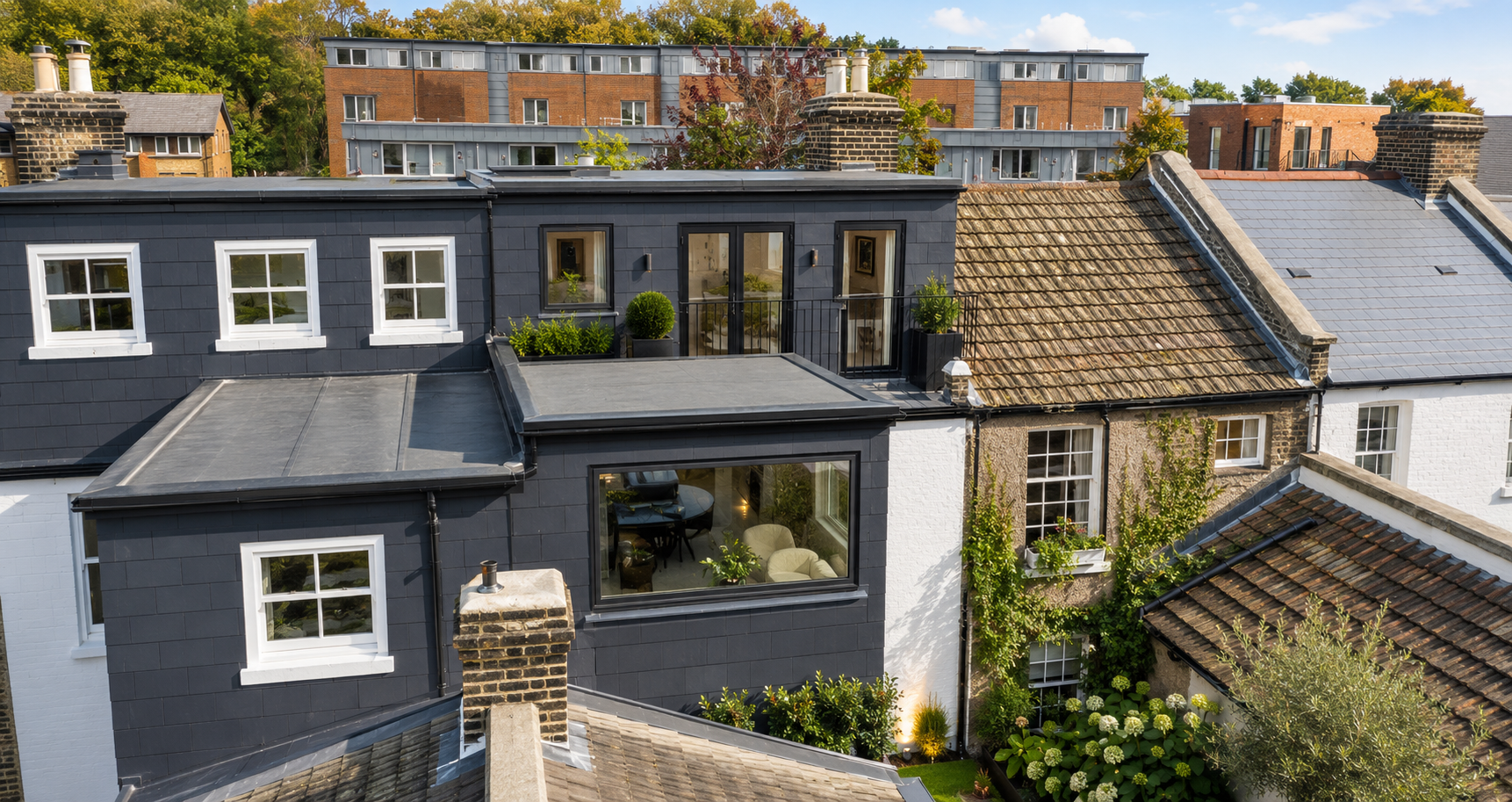 Aerial view of a modern, black-painted rooftop apartment with large windows, plants, and patio door, surrounded by traditional houses with brick and tile roofs, in a neighborhood with trees and apartment buildings.