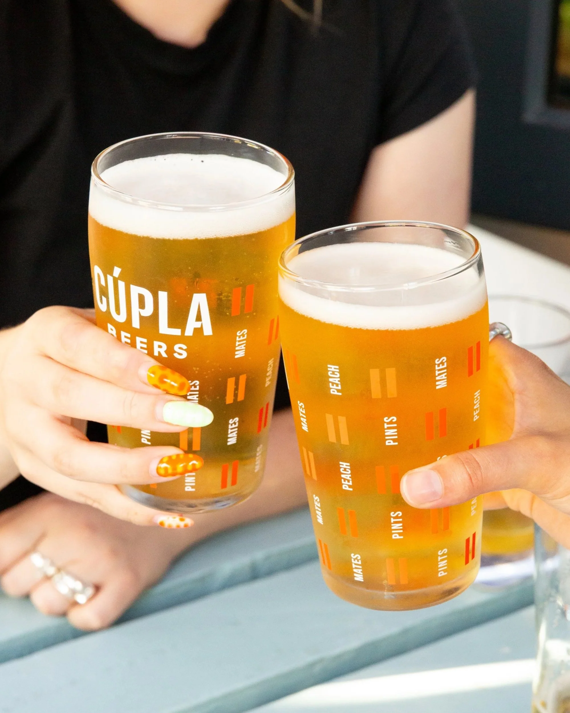 Cúpla Beers Branded Pint Glass with Logo, Dual Line Motif, Mates, Pints and Peach text also on glass. Glasses being toasted between two people.
