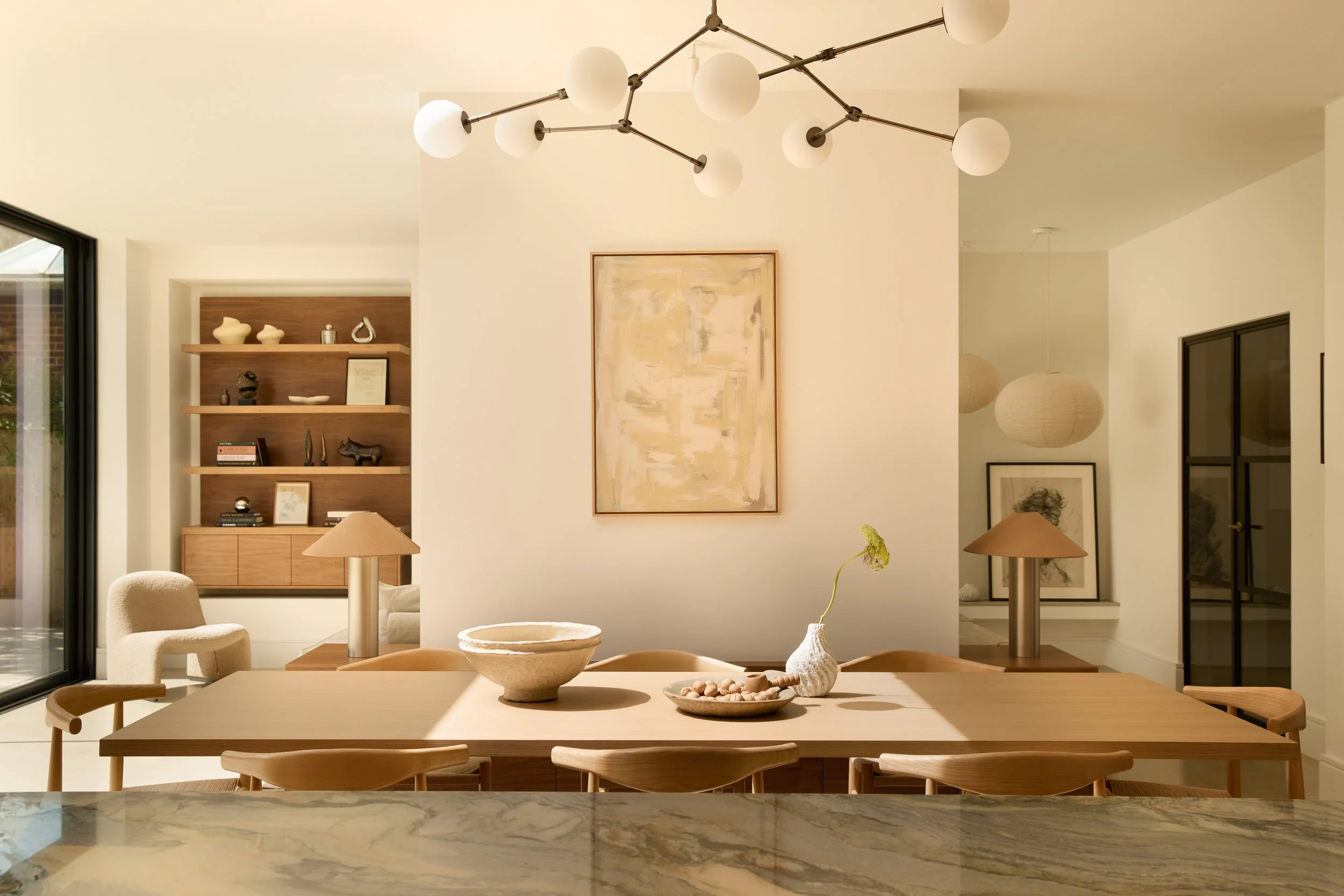 A modern dining room with a light wooden table and matching chairs, decorated with bowls and small vases. A large abstract painting hangs on a white wall, with decorative shelves and artwork nearby. There are table lamps on side tables, and a large window allows natural light into the space. The ceiling features a contemporary chandelier with multiple globes.
