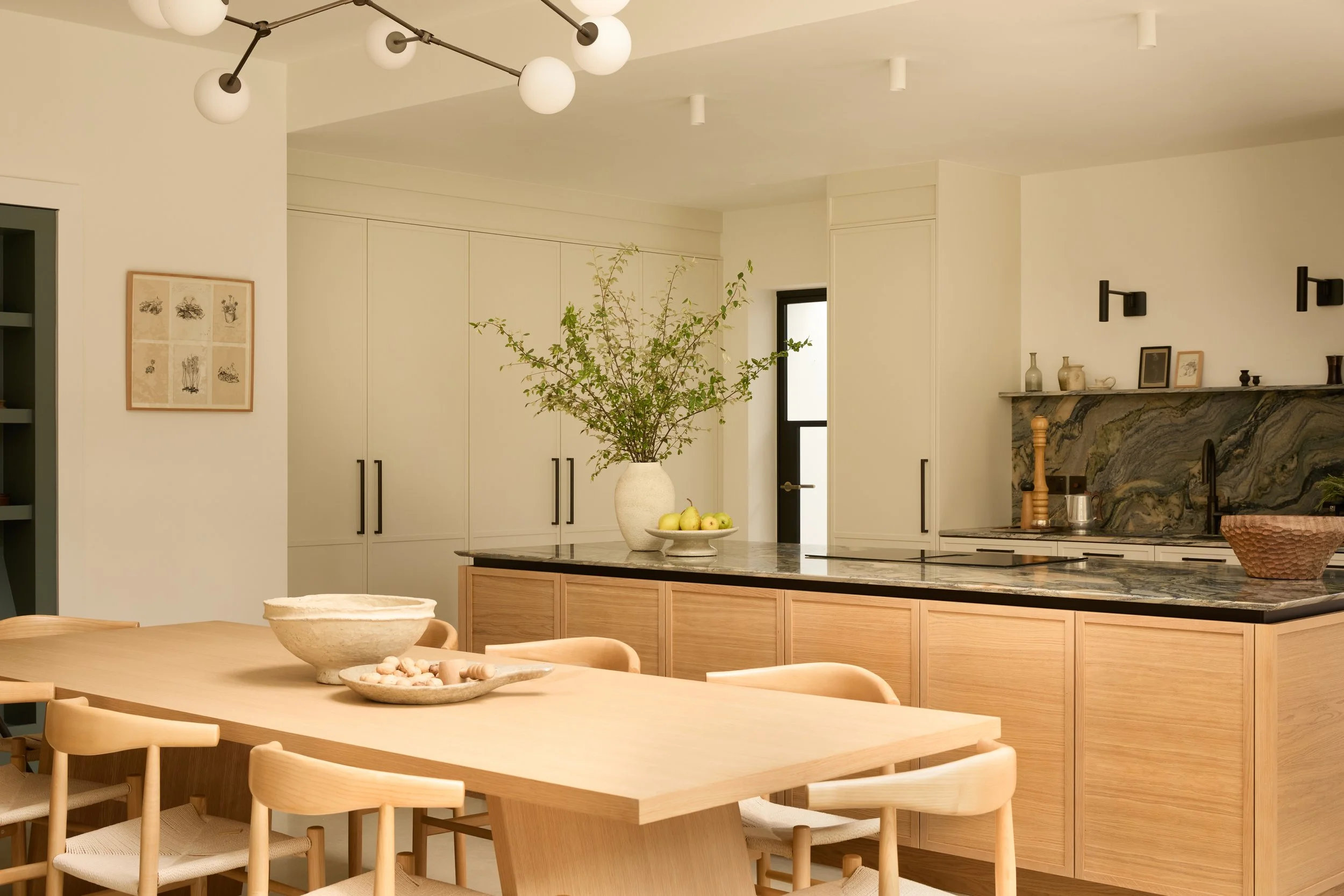 A modern kitchen with a large wooden dining table, surrounded by light wood chairs, a central island with a granite countertop, a large plant in a vase, and decorative items on a shelf above a marble fireplace.