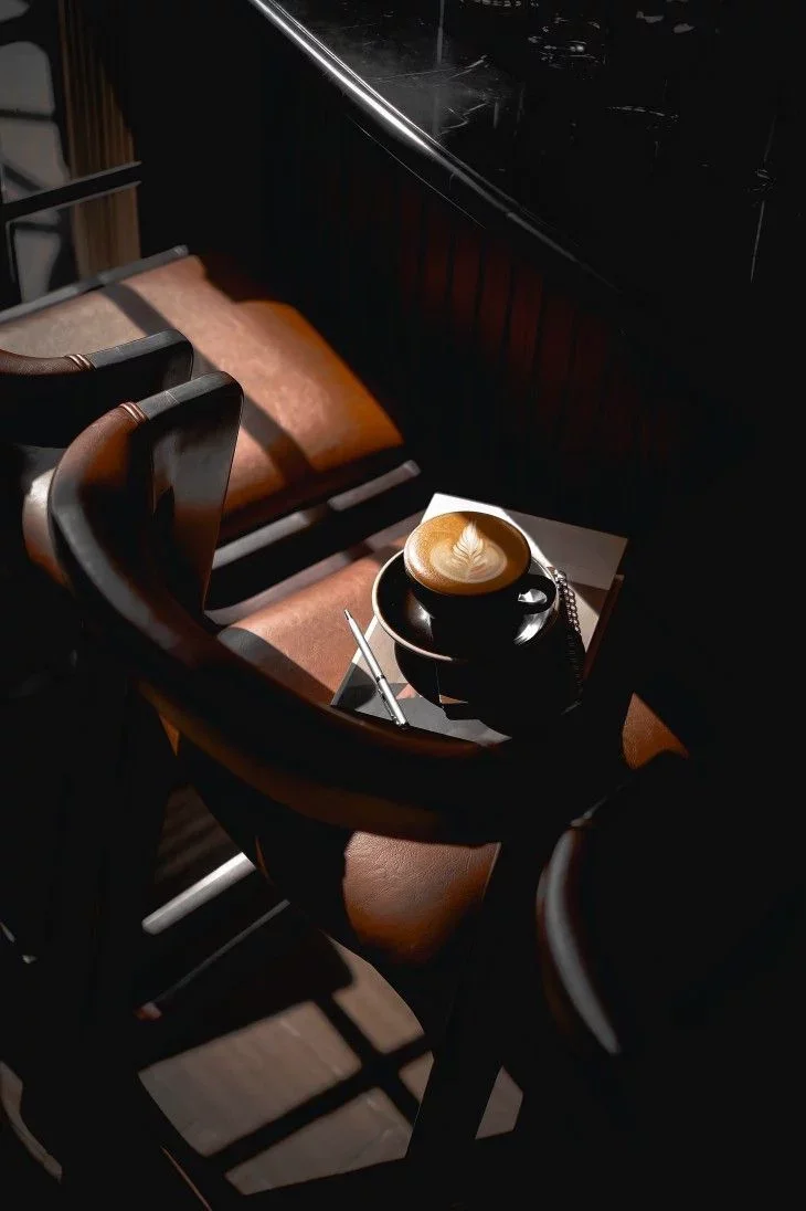 A black coffee cup with latte art on top sits on a small black table beside brown chairs with dark wood and leather cushions, next to a black countertop, with sunlight casting shadows.