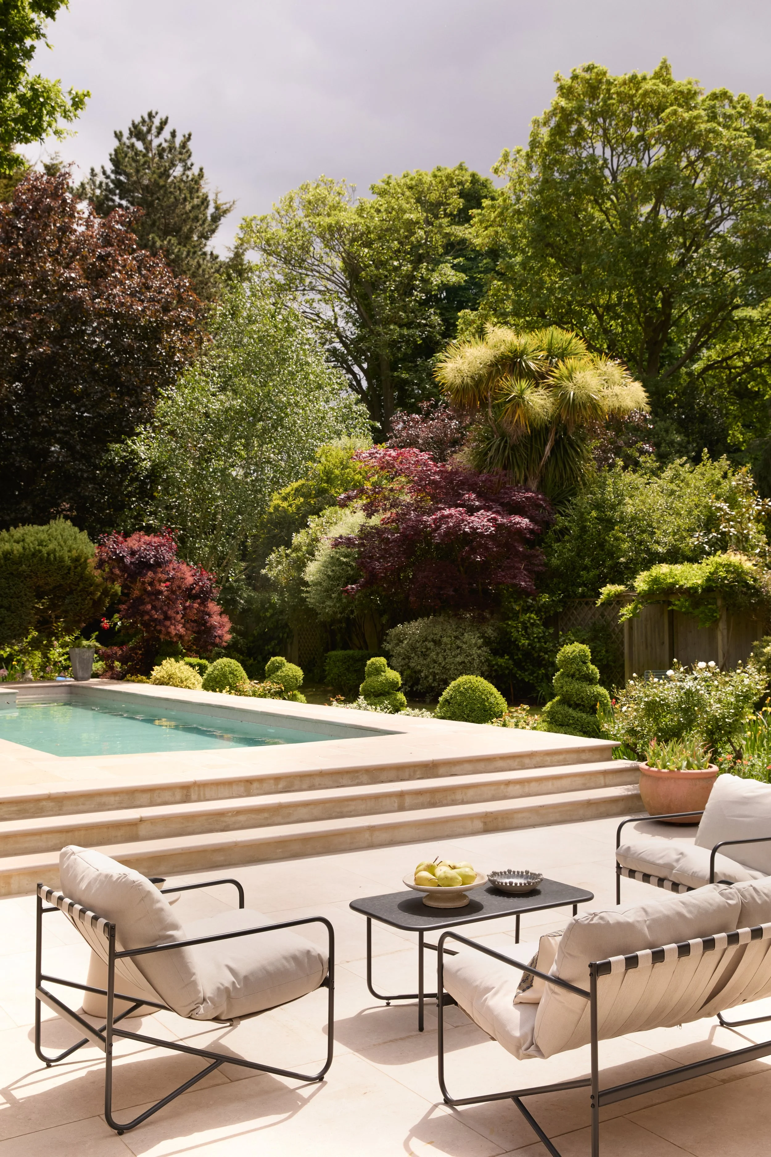 Outdoor patio with cushioned chairs, a table with a bowl of green apples, and a swimming pool, surrounded by lush greenery and trees, under cloudy sky.