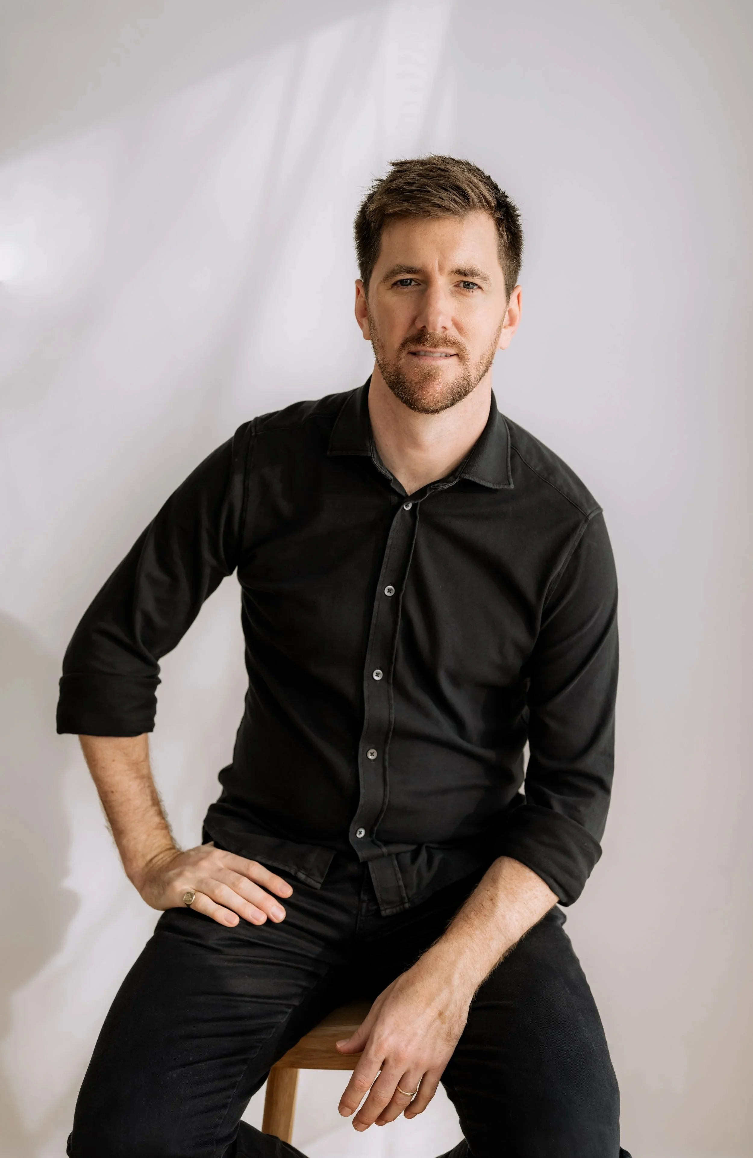 A man with short dark hair and a beard, wearing a black button-up shirt, sitting on a wooden stool with one hand on his thigh and the other on his waist, against a plain white background.