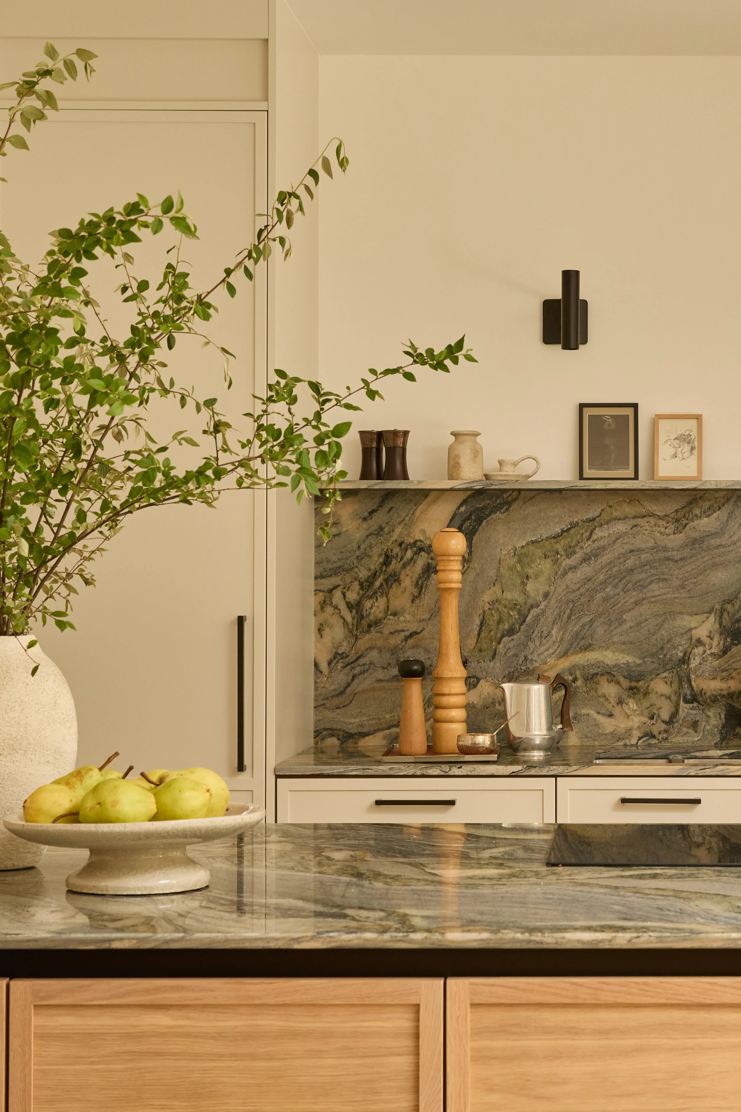 A kitchen with a marble counter, displaying a bowl of pears, potted plants, and decorative items on a marble backsplash. There are framed pictures and a black wall lamp on a beige wall.