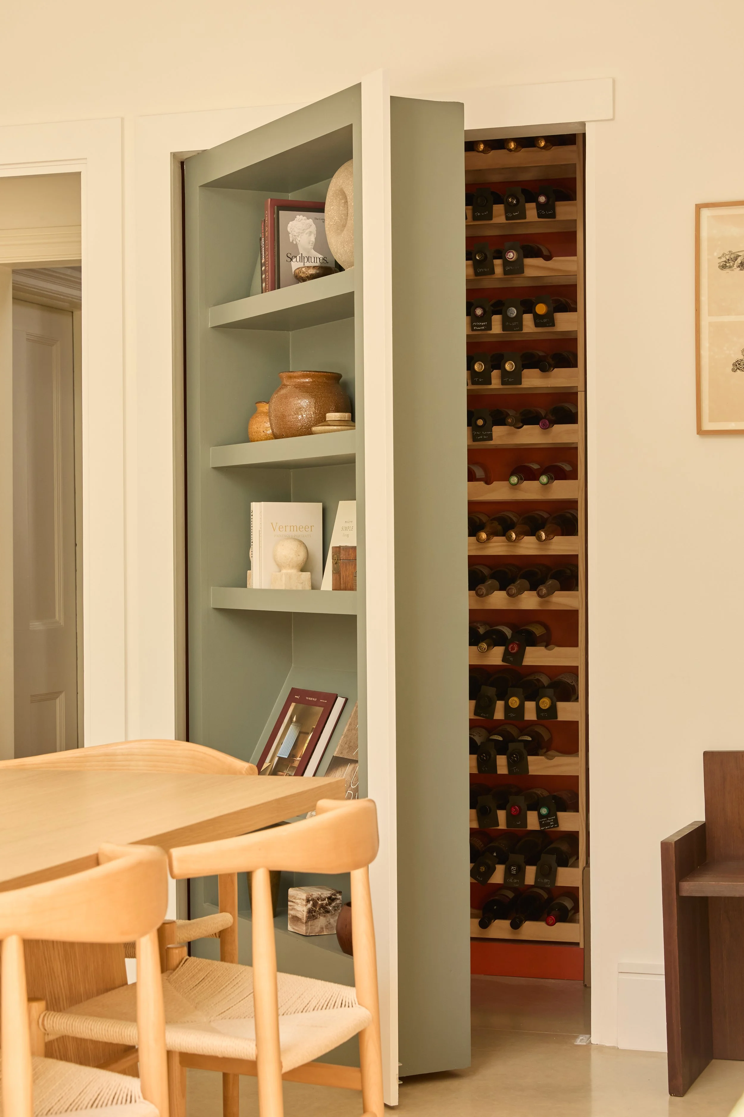 A wooden dining table with light-colored chairs in a room, with a green bookshelf containing decorative items and books, and a wine rack filled with wine bottles in the background.