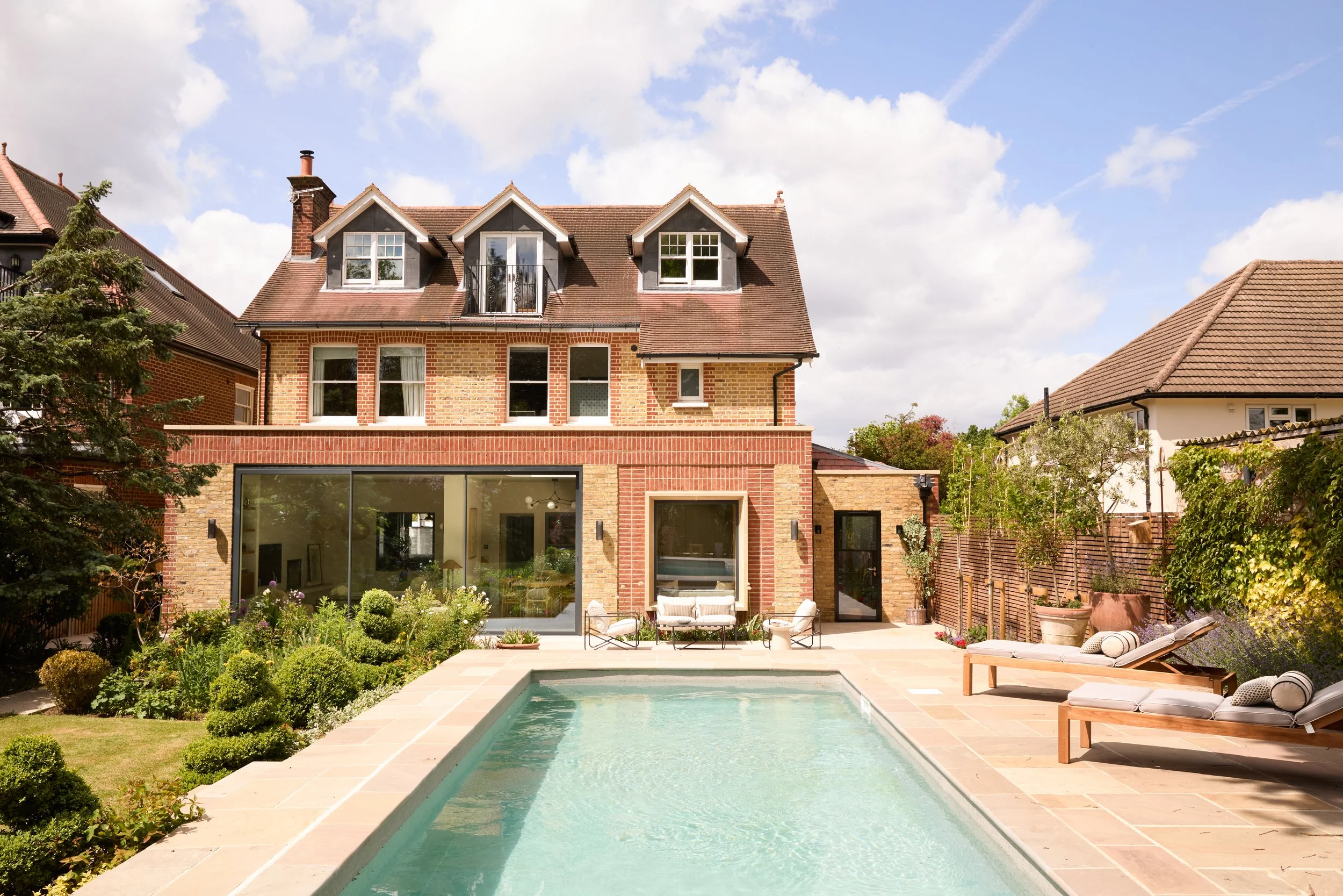 A backyard with a swimming pool surrounded by a patio area, lounge chairs, and outdoor furniture. There are lush plants and trees, with a brick house featuring multiple windows and a balcony. The house has a gabled roof, and the sky is blue with a few clouds.