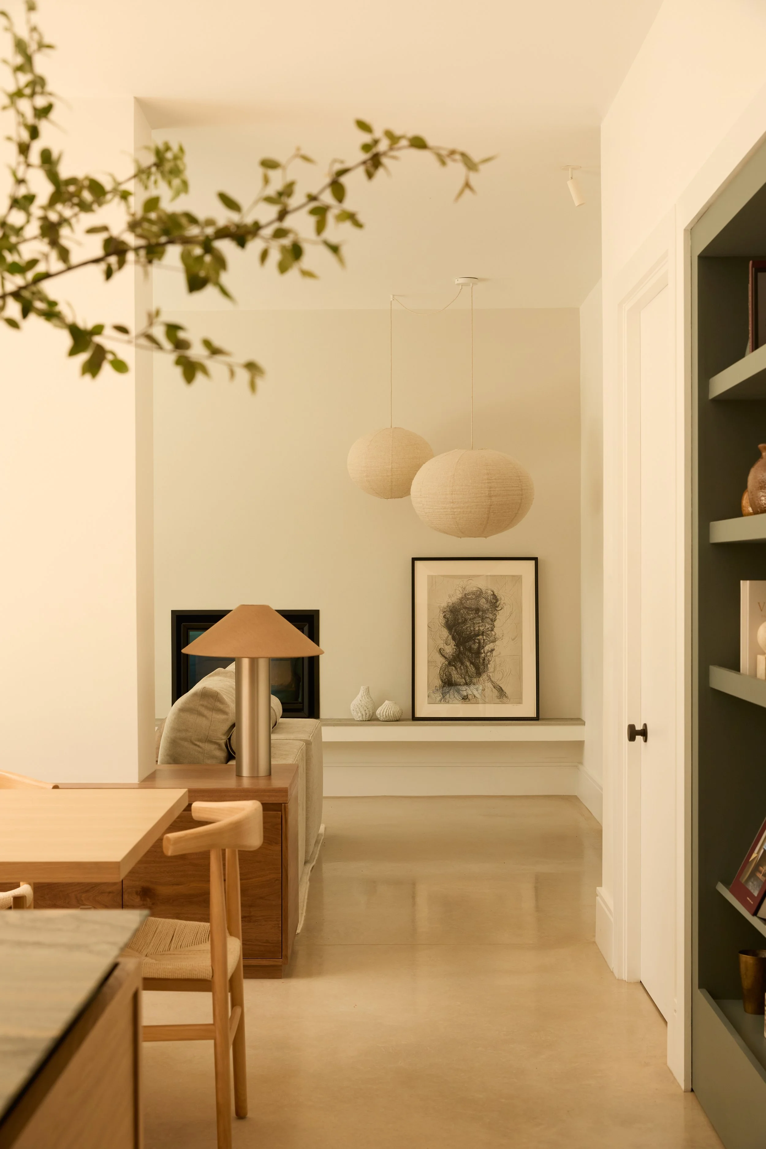 A cozy, modern interior room with beige walls, hanging spherical pendant lights, framed artwork, a beige couch, a wooden table with chairs, and a bookshelf with decorative items.