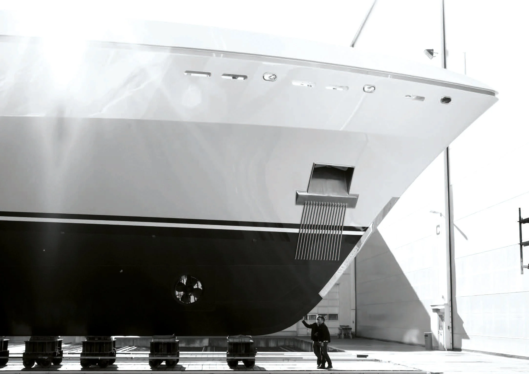 A large ship is under construction or maintenance indoors with a person standing nearby pointing at it. The ship's hull is predominantly white and black, with some structural openings and ventilation grilles visible.