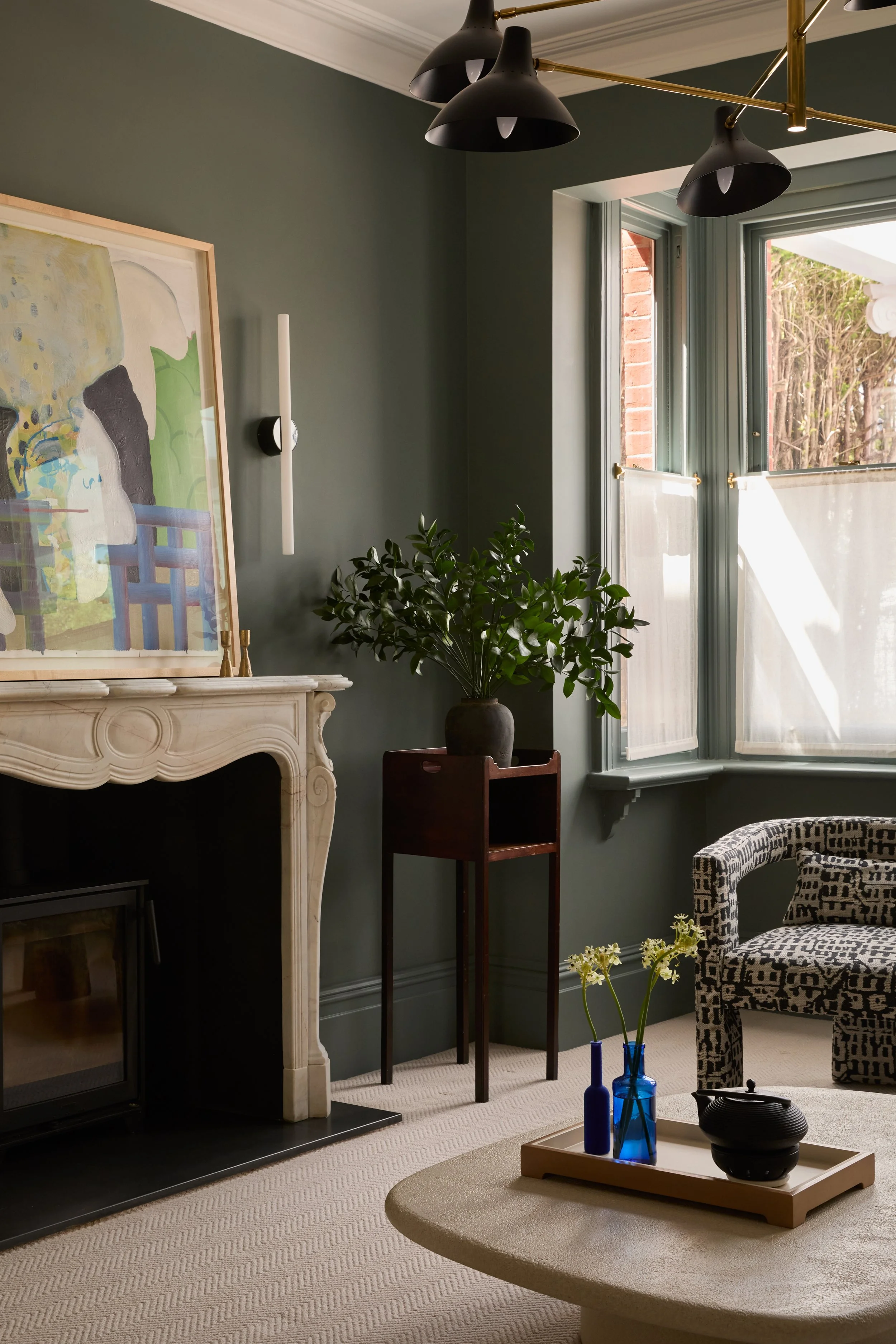 A cozy living room corner with a fireplace, green walls, a large abstract painting, a window letting in natural light, a potted plant, a small red table, and a patterned armchair, accented with blue vases and a tray on a round table.