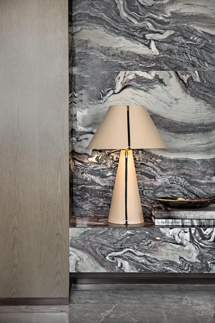 A marble wall with black, white, and gray swirling patterns, a beige lamp with a split shade, and a stack of books on a marble shelf with a small decorative boat on top.