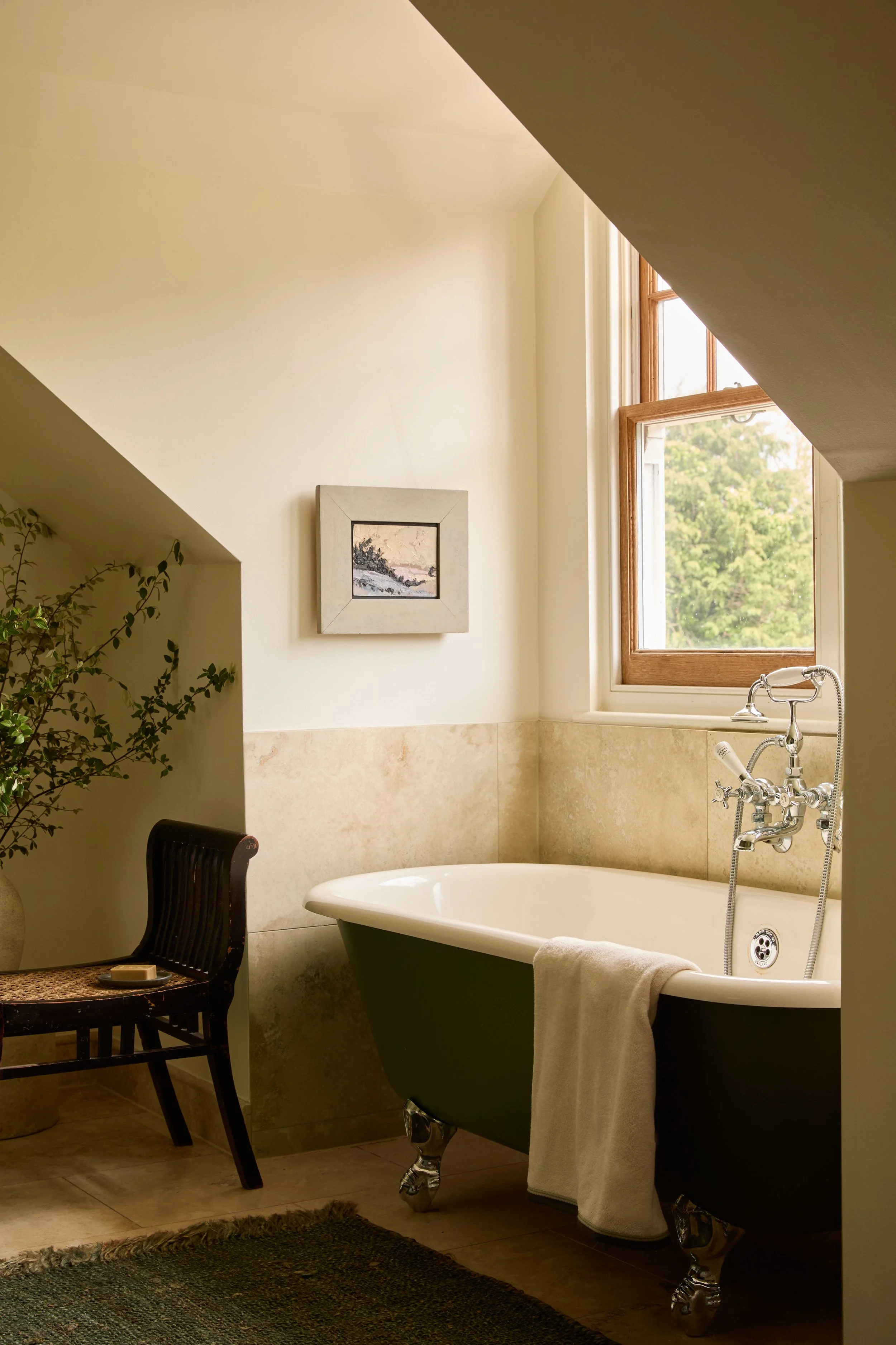 A cozy bathroom with a vintage clawfoot bathtub, a window with wooden framing, a framed picture on the wall, a dark wooden chair, a small table, and a plant in the corner.