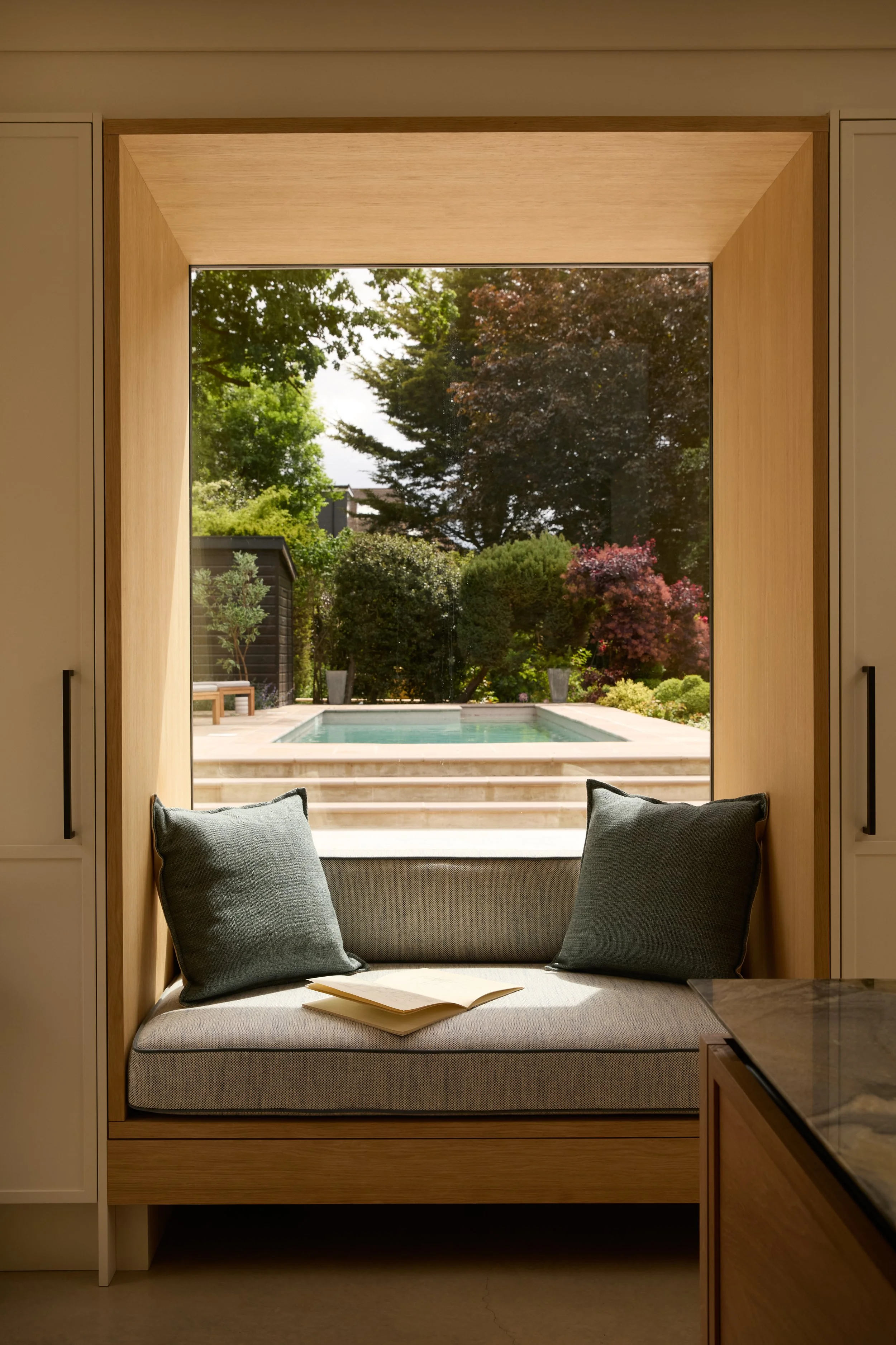 View through a large window showing a backyard with trees, a pool, and a sitting area. Inside, there is a cushioned window seat with two dark grey pillows and an open book.