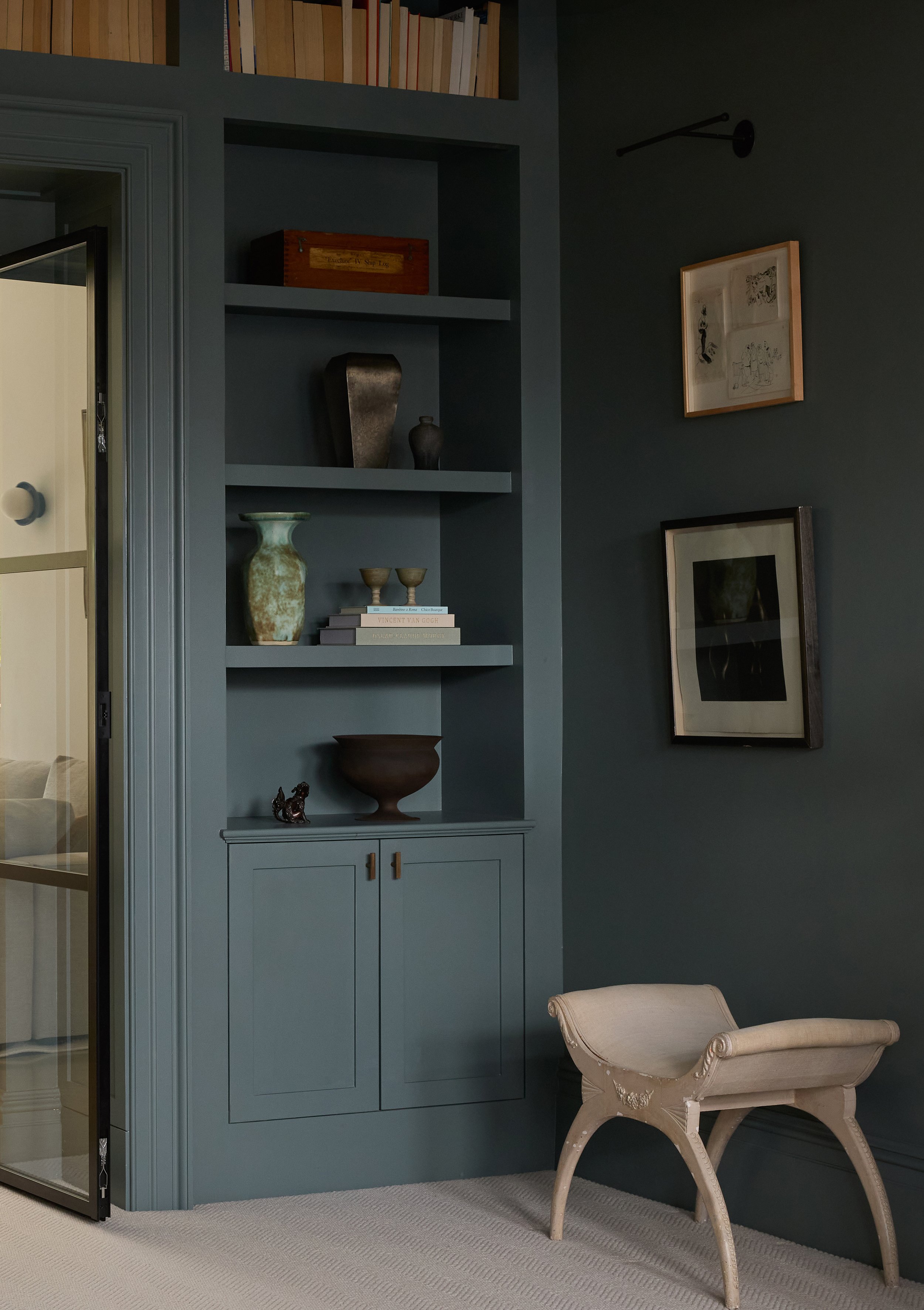 A built-in shelving unit with books and decorative items, a framed artwork on the wall, a beige upholstered bench, and part of a glass door in a room with dark teal walls and light-colored carpet.