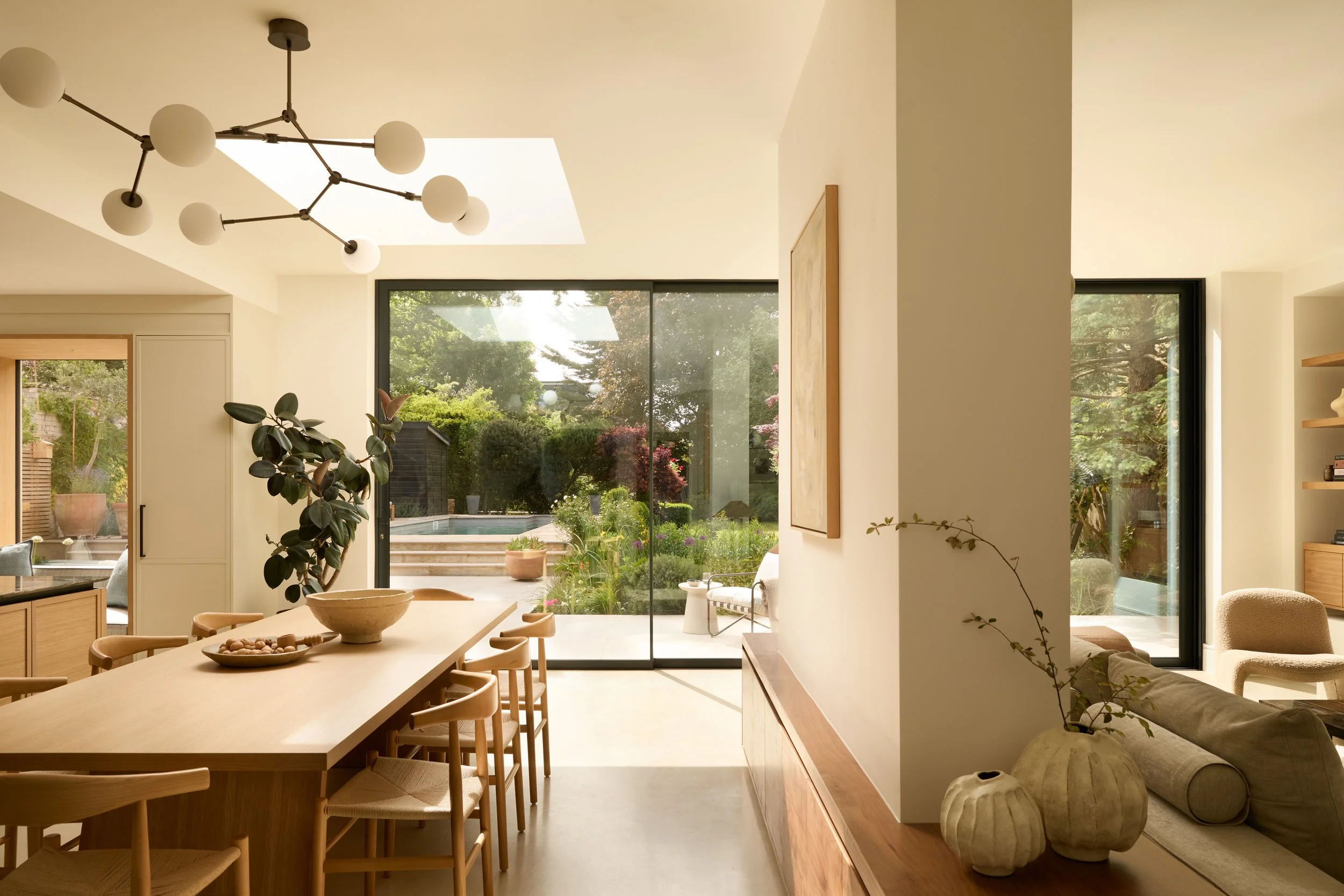 A modern dining and living area with large sliding glass doors leading to a backyard with a swimming pool and garden. The space features a wooden dining table with chairs, a lounge area with beige furniture, and decorative plants.