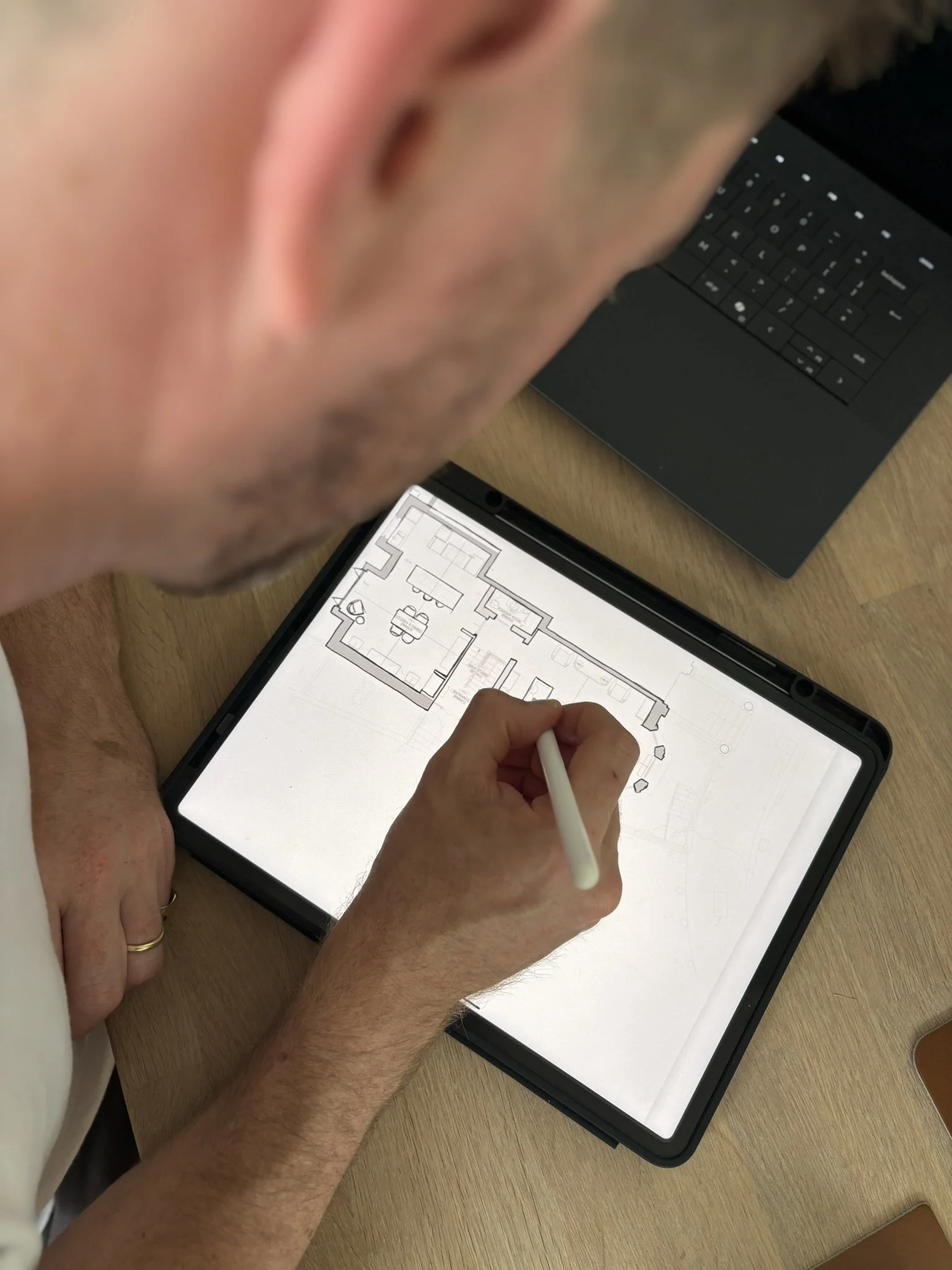 A person is drawing a floor plan on a tablet with a stylus. There is a laptop on the table next to them.