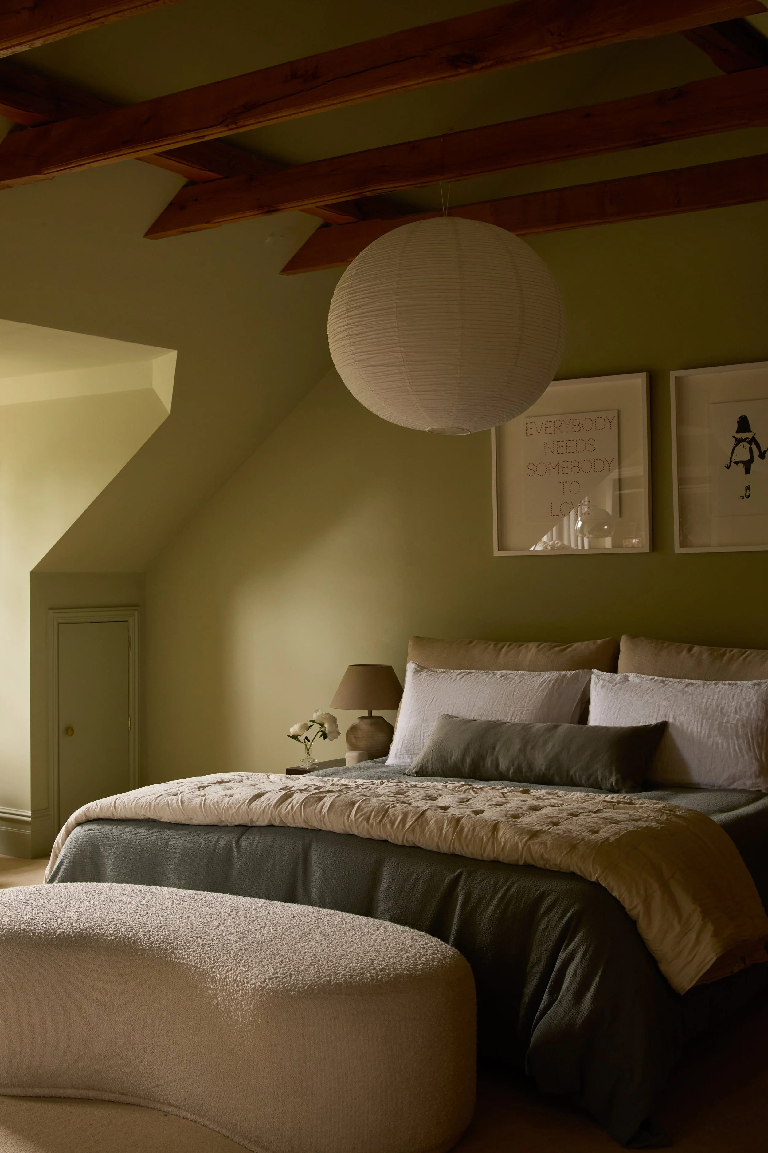 A cozy bedroom with a bed, decorative pillows, a nightstand with a lamp and flowers, framed artwork on the wall, a quilted bench at the foot of the bed, and a paper lantern ceiling light.