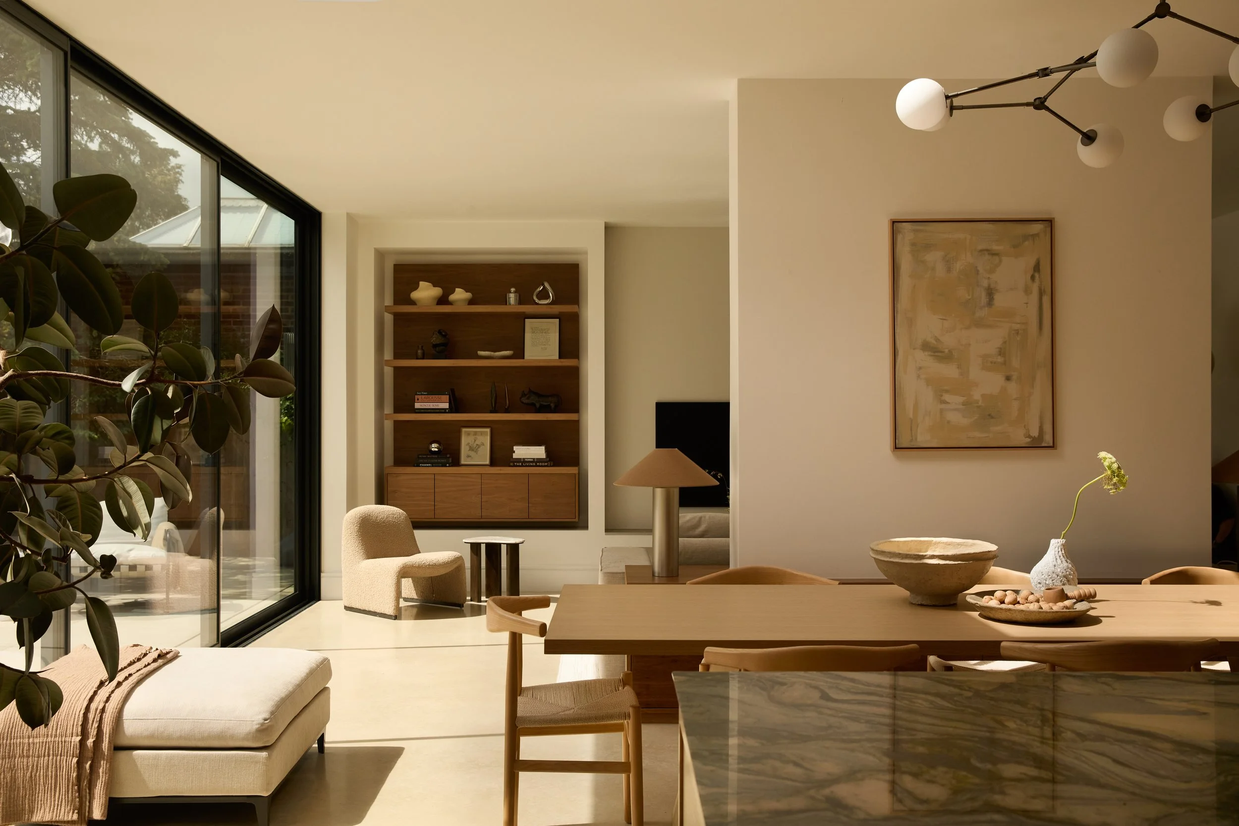 Living room with large glass sliding doors, wooden shelving with decorative items, beige armchair, modern chandelier with white globes, large abstract painting, marble table, and minimalistic decor.