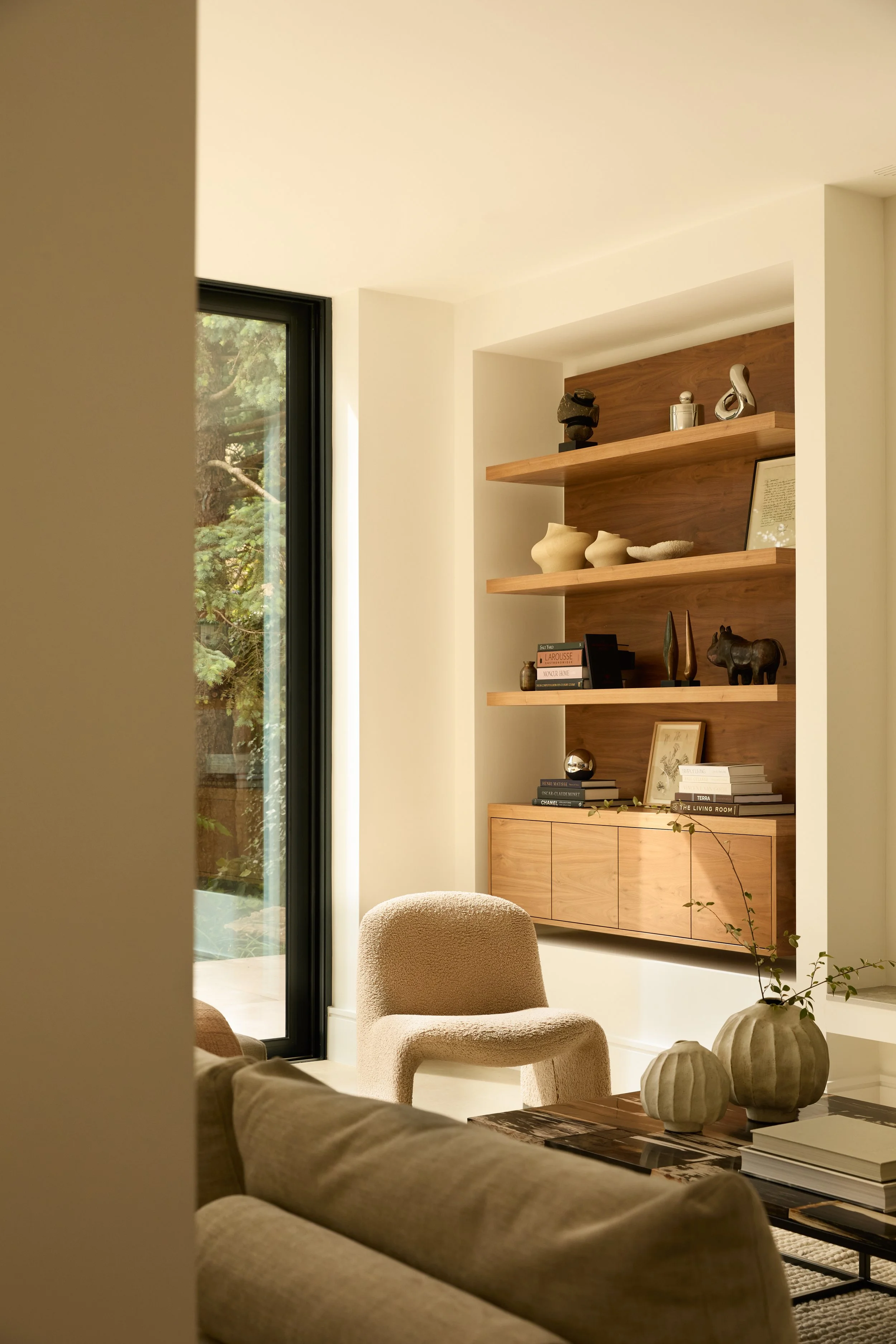 A living room with beige chairs, a wooden sideboard, floating shelves with decorative objects, and a window showing greenery outside.