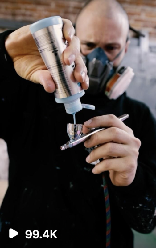 A person wearing a respirator mask spray painting a small object under studio lighting.