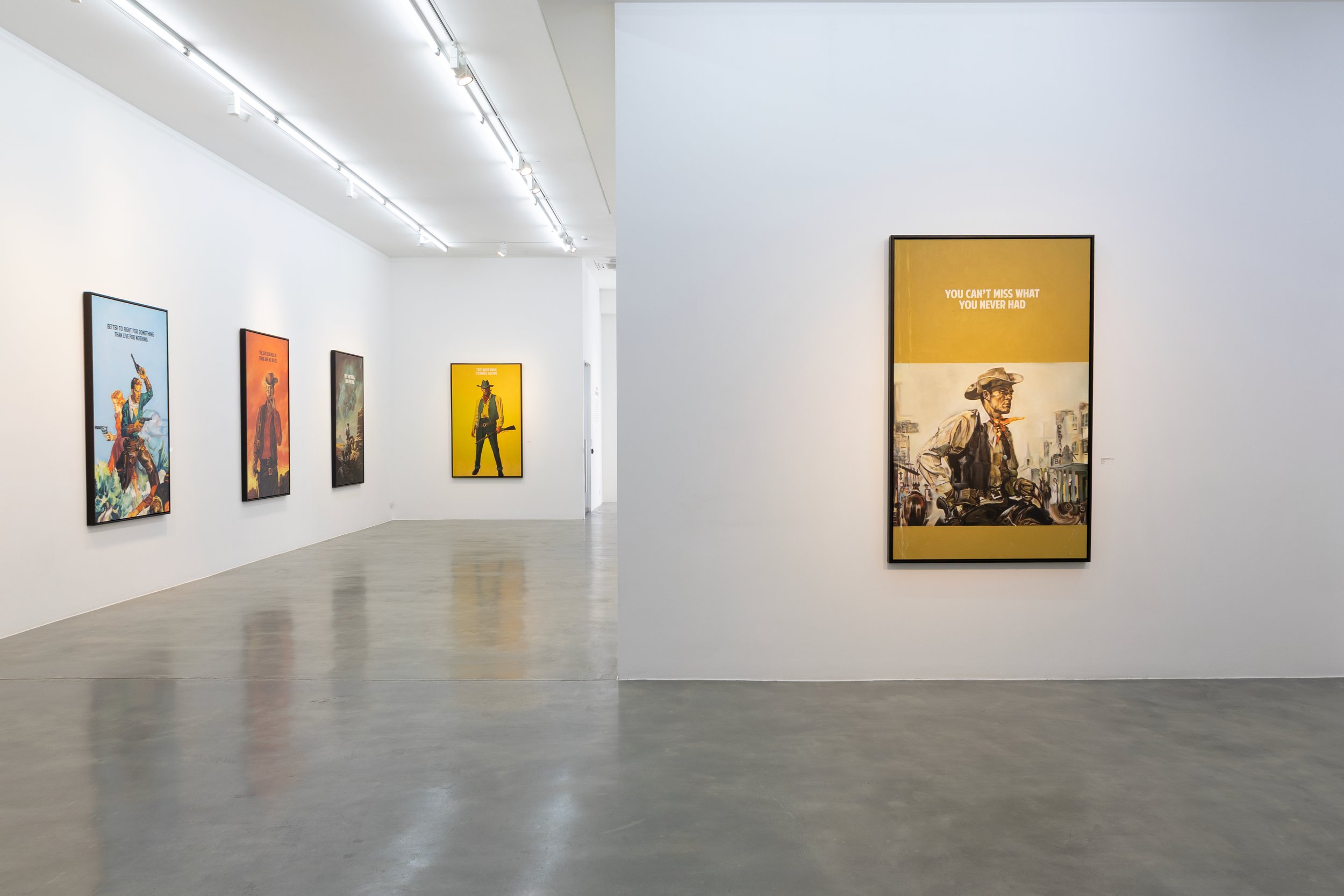 An art gallery with paintings displayed on white walls, featuring Western-themed artworks including cowboys and signage, with a polished concrete floor and bright overhead lighting.