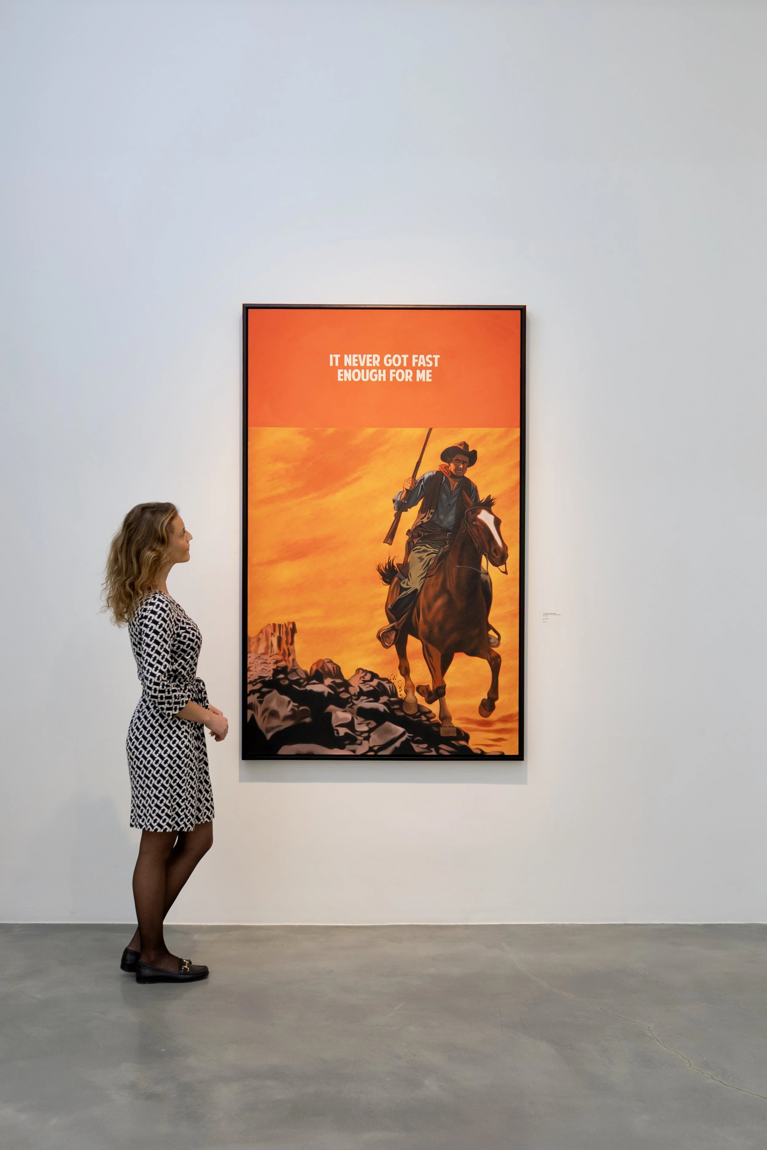 Woman with curly hair, wearing black tights, black shoes, and a patterned dress, observing a large painting of a cowboy riding a horse against a sunset background.