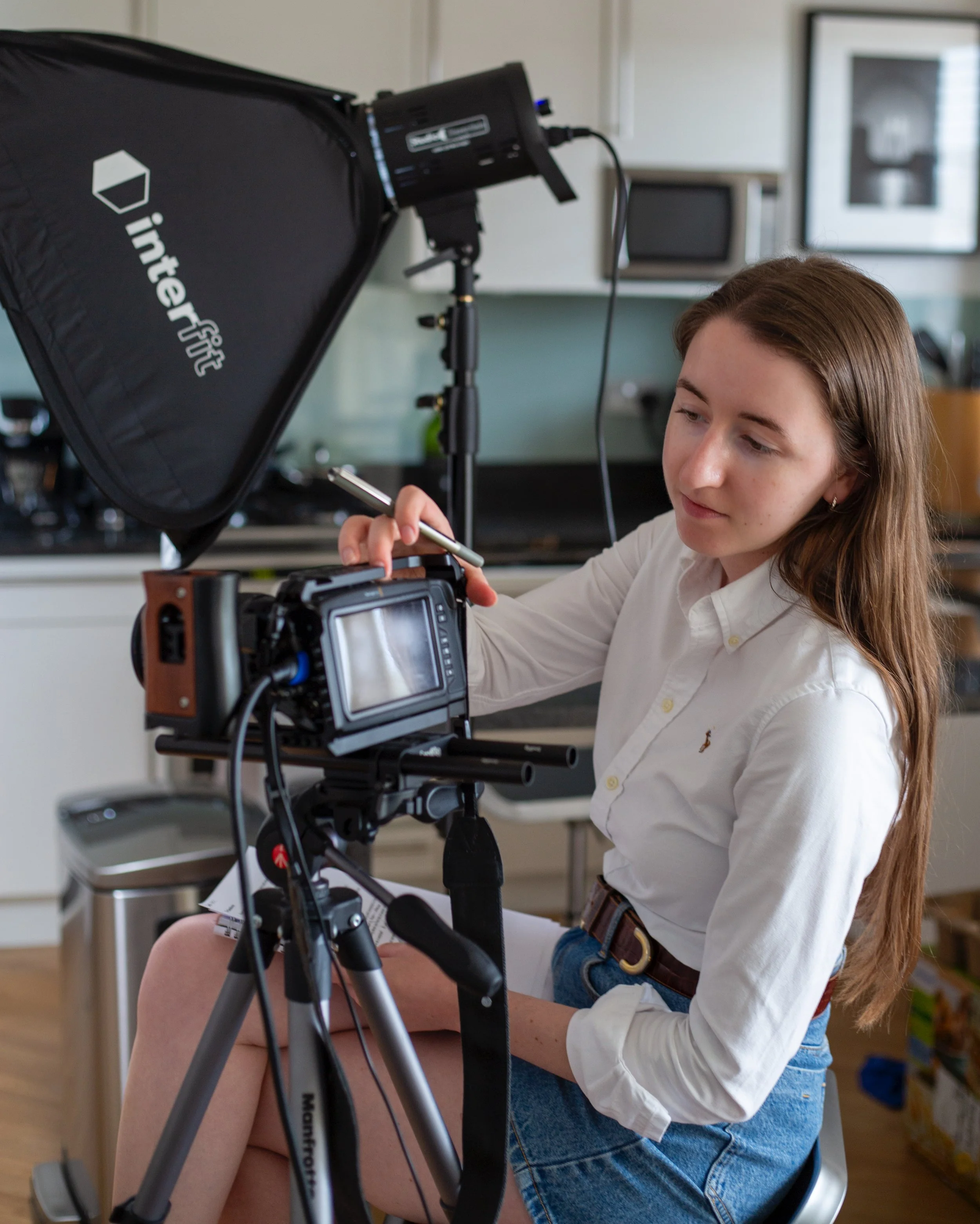 A woman with long brown hair, wearing a white shirt and blue jeans, is adjusting a professional camera on a tripod. A large softbox light with the logo 'interfit' is illuminating the scene. The background shows kitchen cabinets and a microwave.