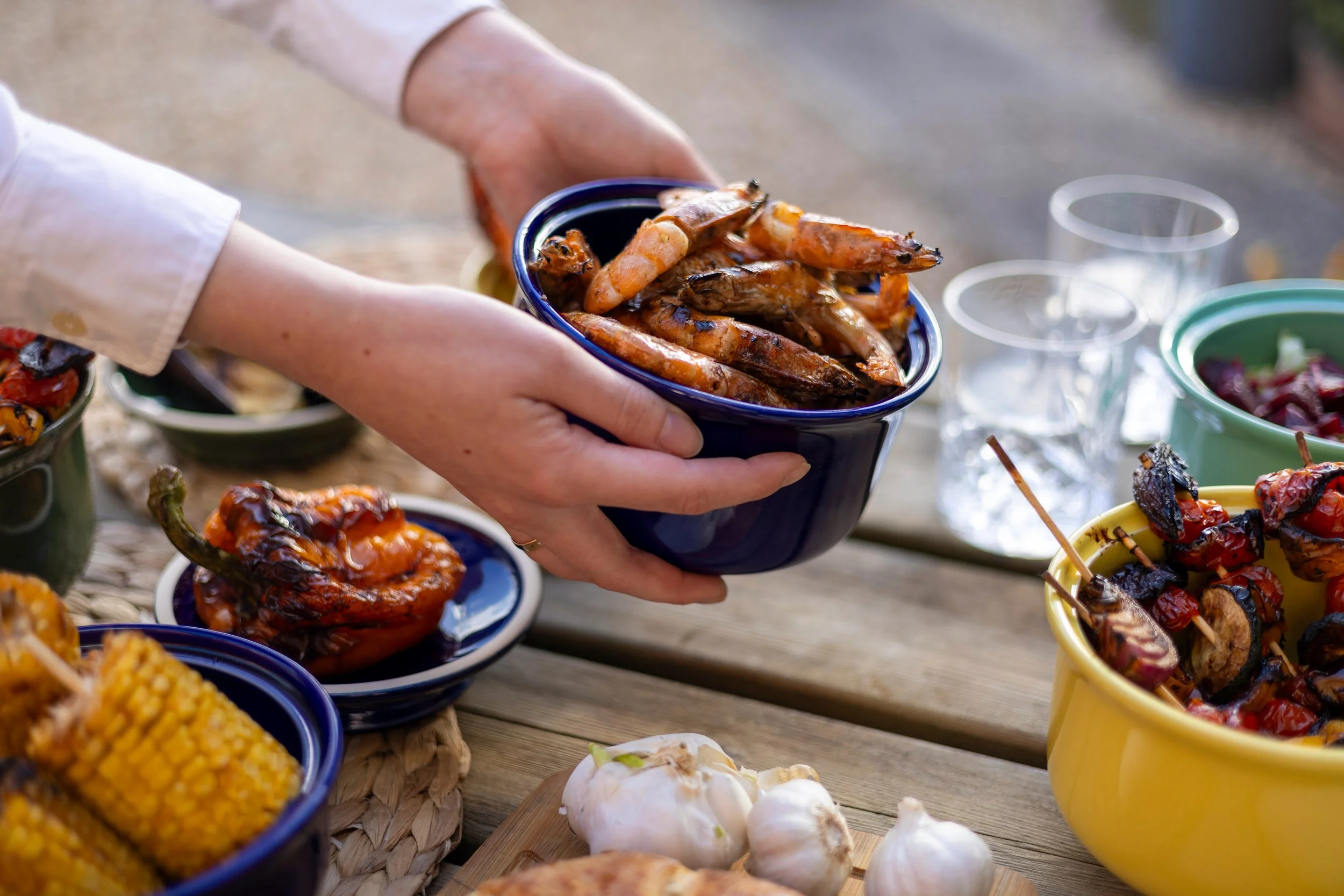Person holding a bowl of grilled shrimp at an outdoor food gathering with various dishes on a wooden table.