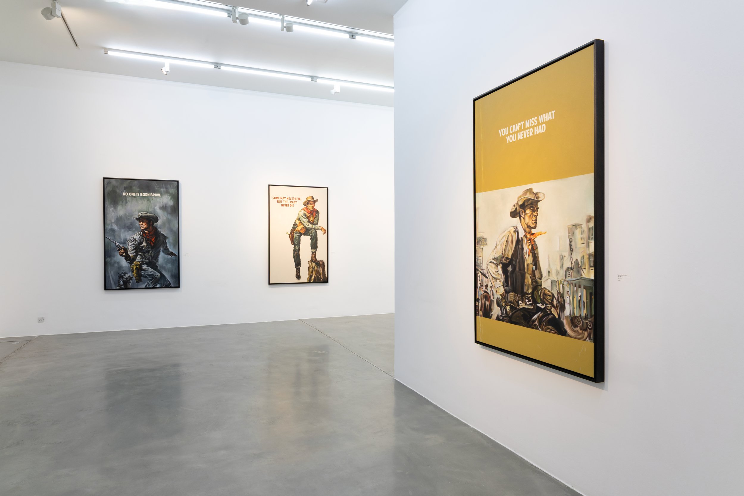 Art gallery with framed paintings of cowboys and motivational quotes displayed on white walls, fluorescent lighting, and concrete flooring.