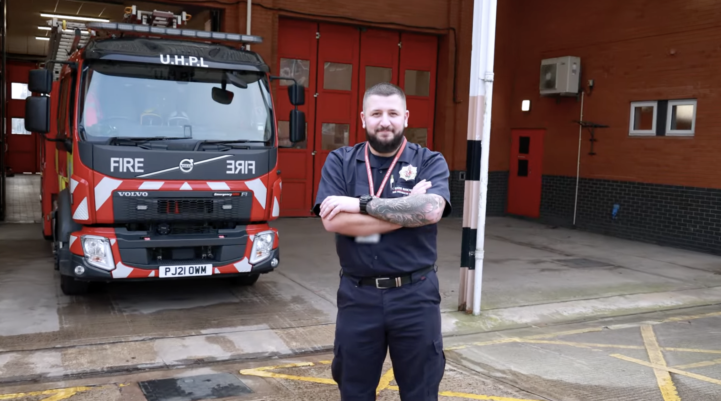 A firefighter with tattoos and a beard standing outside a fire station, crossing his arms, with a fire truck parked behind him.