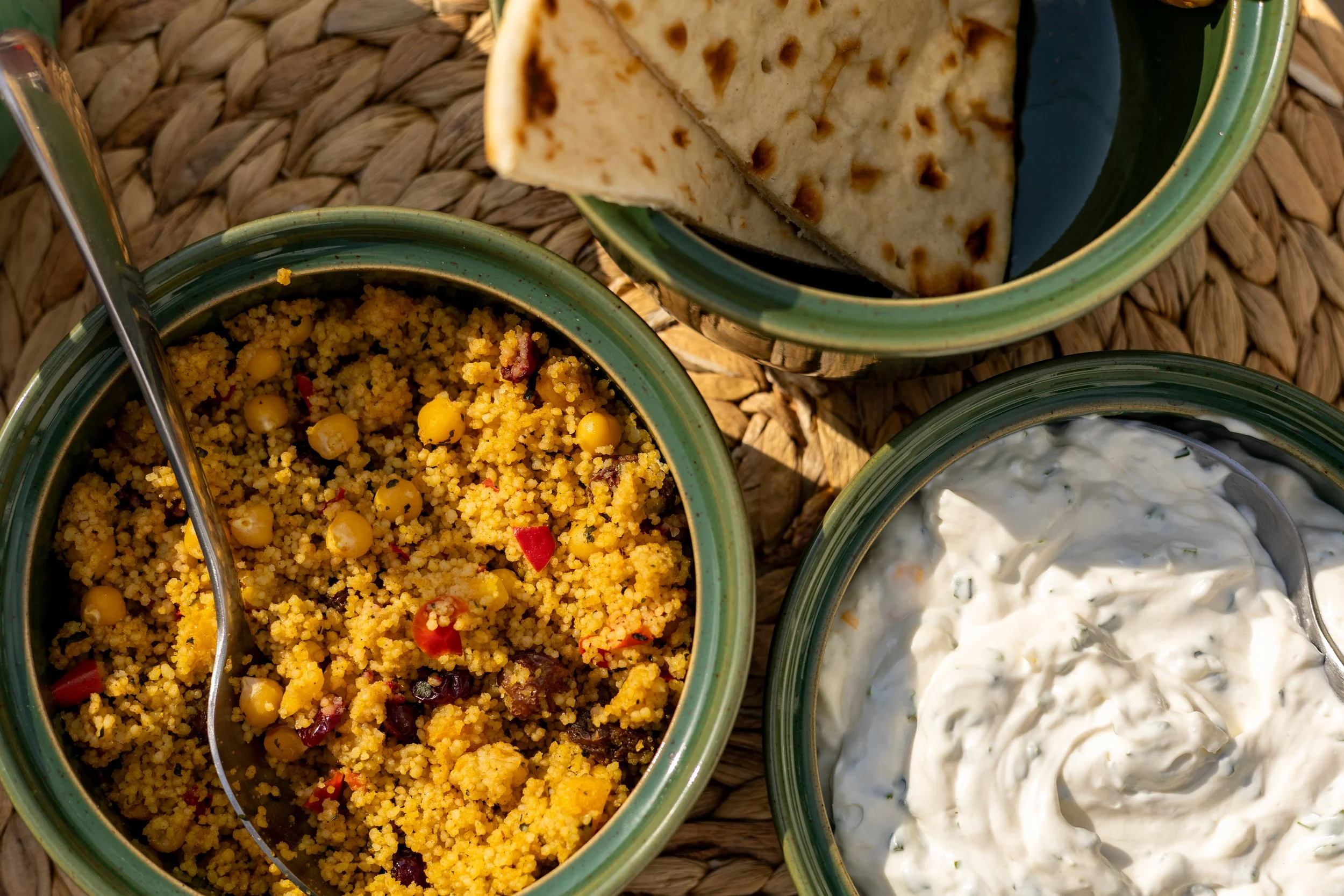 Close-up of three bowls containing Mediterranean dishes. The top bowl has grilled naan bread, the middle bowl contains couscous with chickpeas and vegetables, and the bottom bowl has tzatziki sauce with herbs. The bowls are placed on a woven straw ma