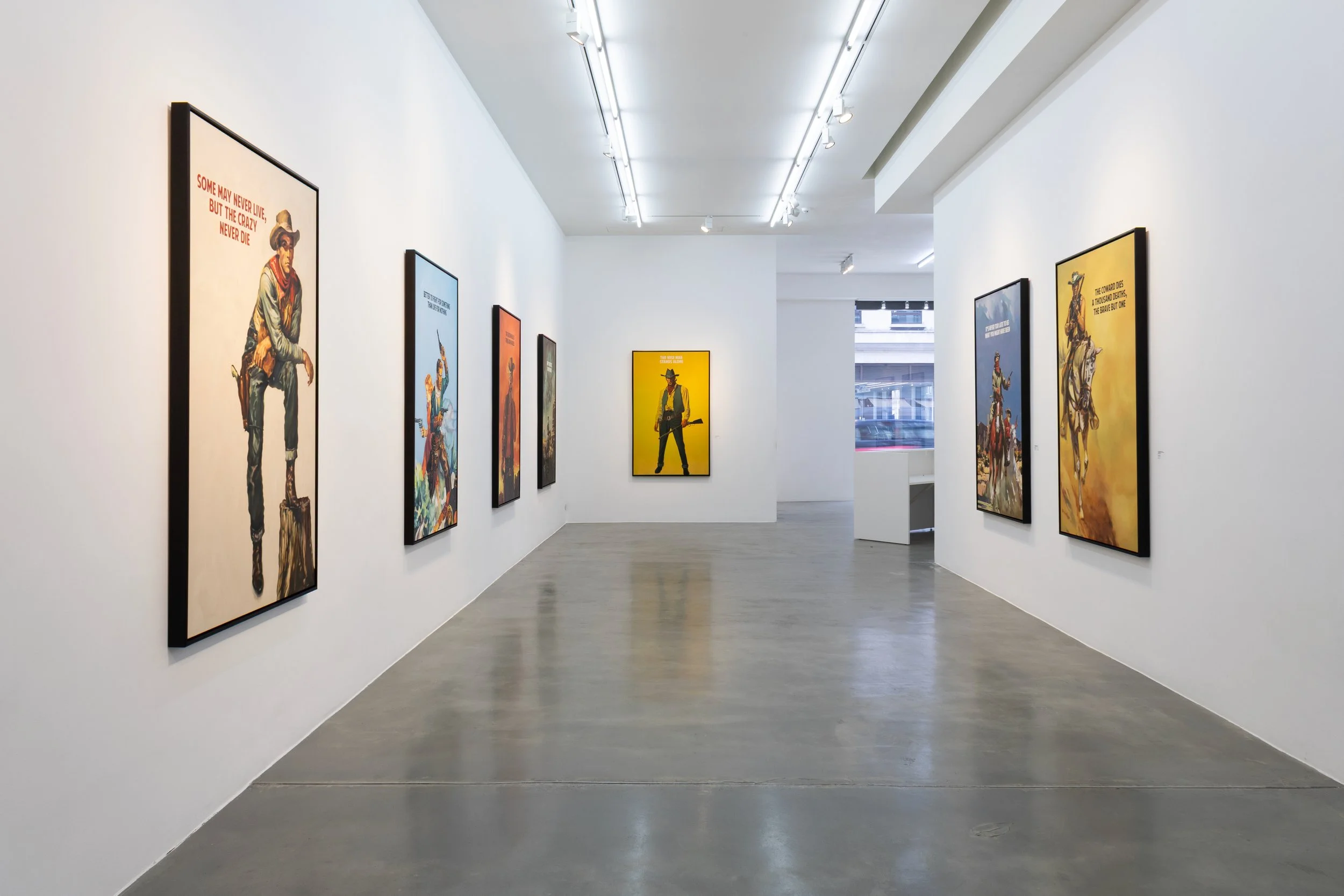 Art gallery with white walls and hardwood floors, featuring multiple framed colorful posters of cowboys in various poses.