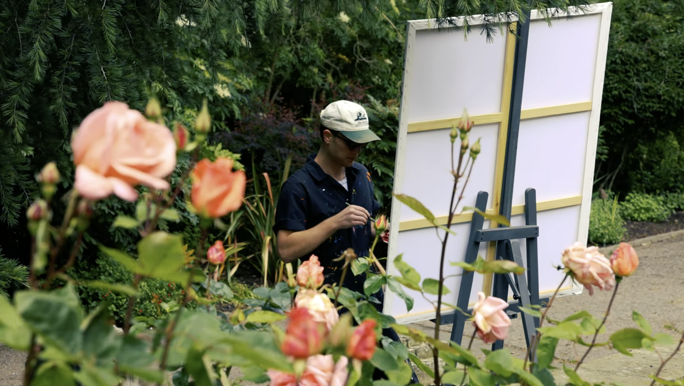 A person wearing a white cap and glasses standing outdoors, painting on a whiteboard with a garden and pink roses in the foreground.