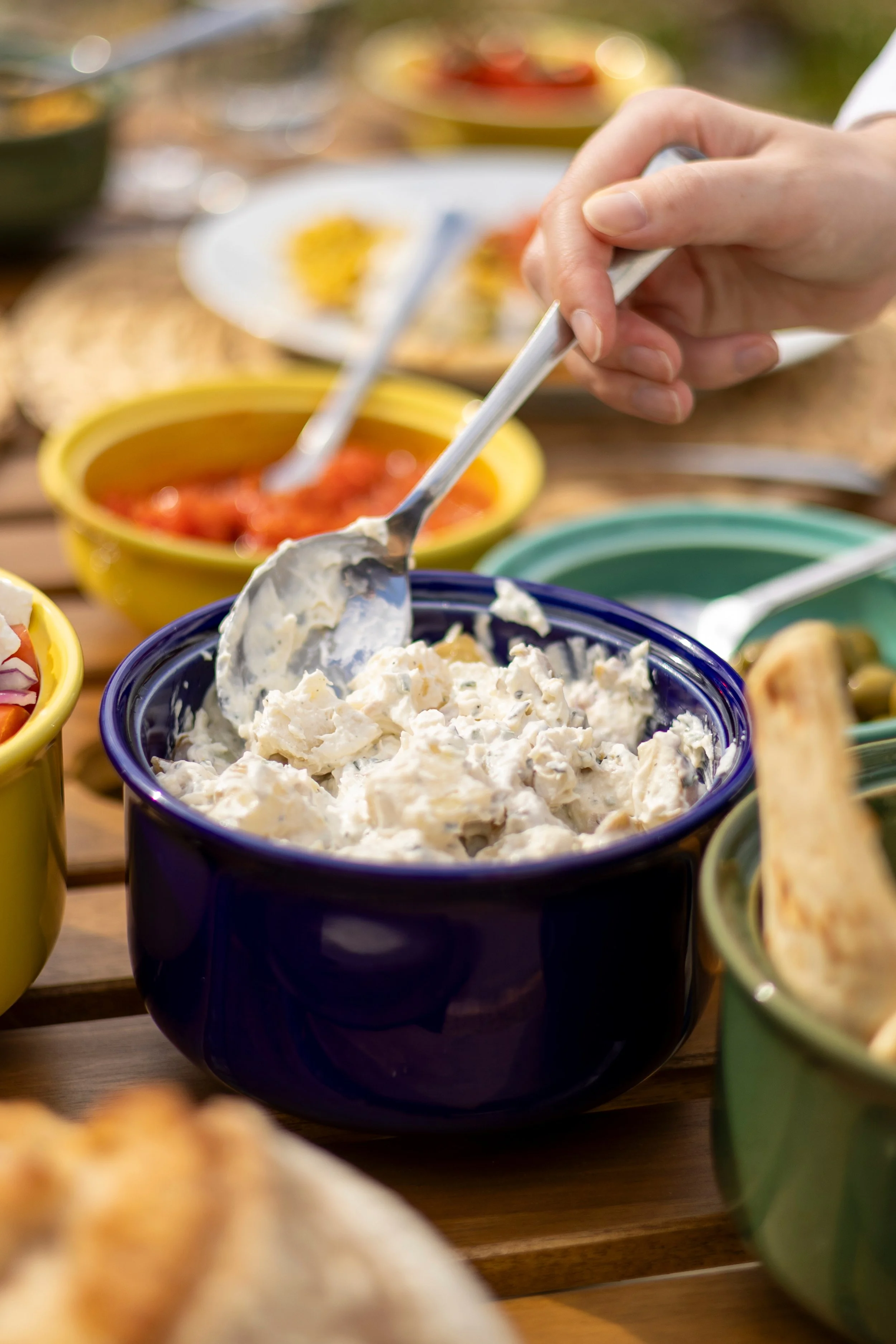 Close-up of a hand scooping creamy salad from a dark blue bowl at a gathering, with various bowls of food on a wooden table.