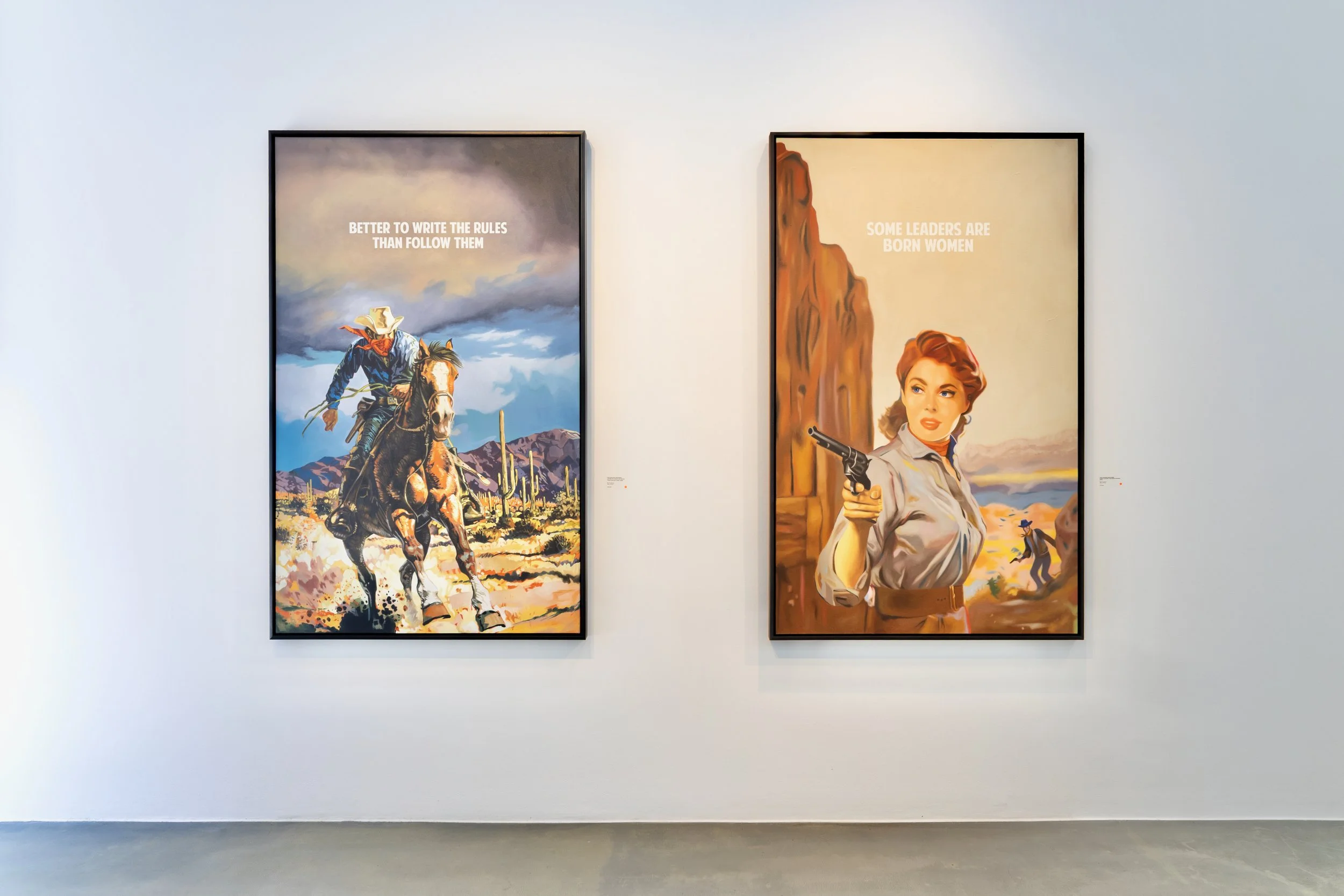 Two framed posters on a white gallery wall, one featuring a cowboy riding a horse with a cloudy sky background and the other showing a woman holding a gun with a desert landscape and rock formation behind her.