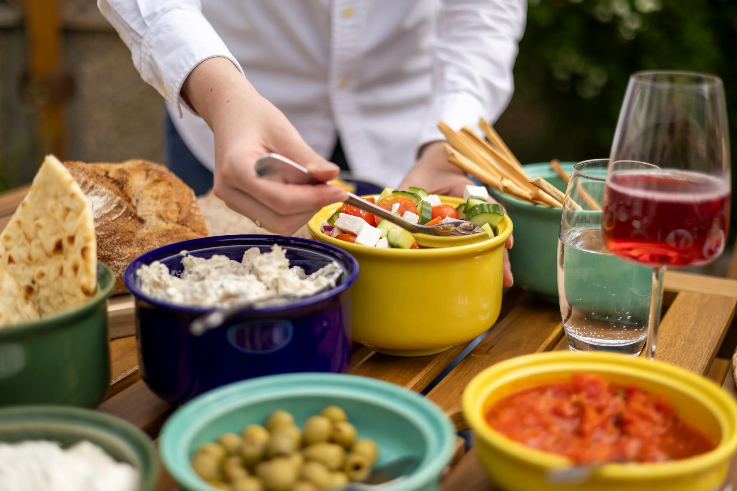 A person preparing a colorful salad with cherry tomatoes, cucumbers, and feta cheese in a yellow bowl, surrounded by various dishes including bread, hummus, and dips, on a wooden table outdoors.