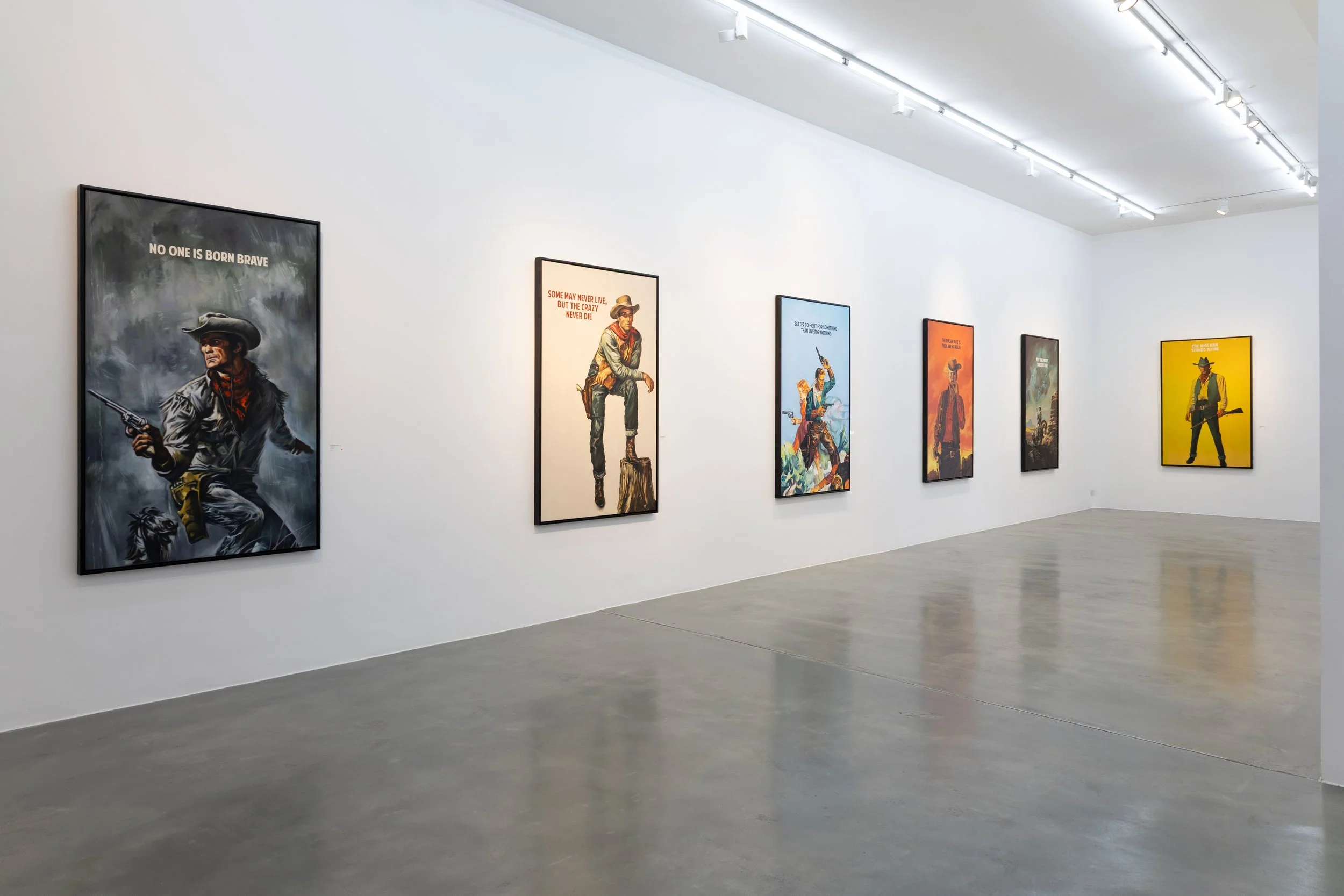 Art exhibition with colorful posters of cowboys and western themes on a white gallery wall, with overhead lighting, in a modern art gallery.