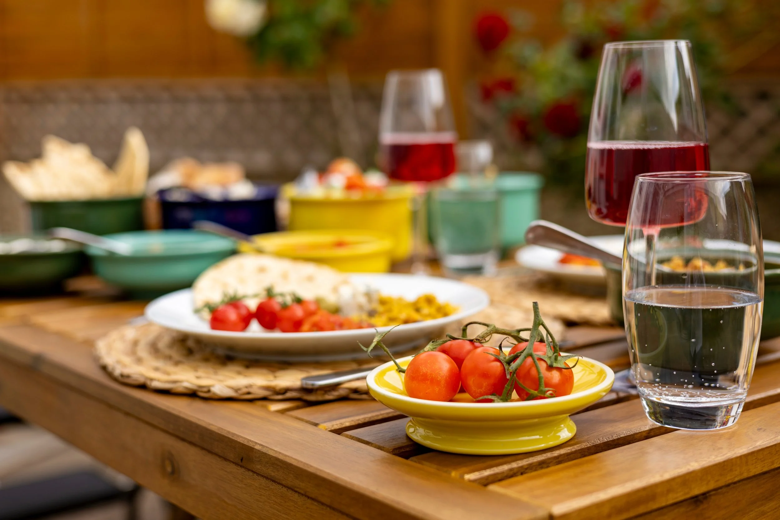 A table set with food and drinks, including a glass of red wine, a glass of water, cherry tomatoes on a yellow plate, and various bowls of snacks, with a blurred background