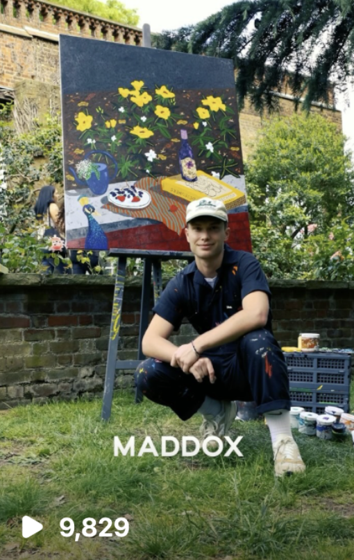 Young man kneeling in front of an outdoor easel with a colorful painting of yellow and white flowers with plants, a basket, a bottle, and a plate of food. There are paint supplies beside him and a garden with trees and bushes in the background.