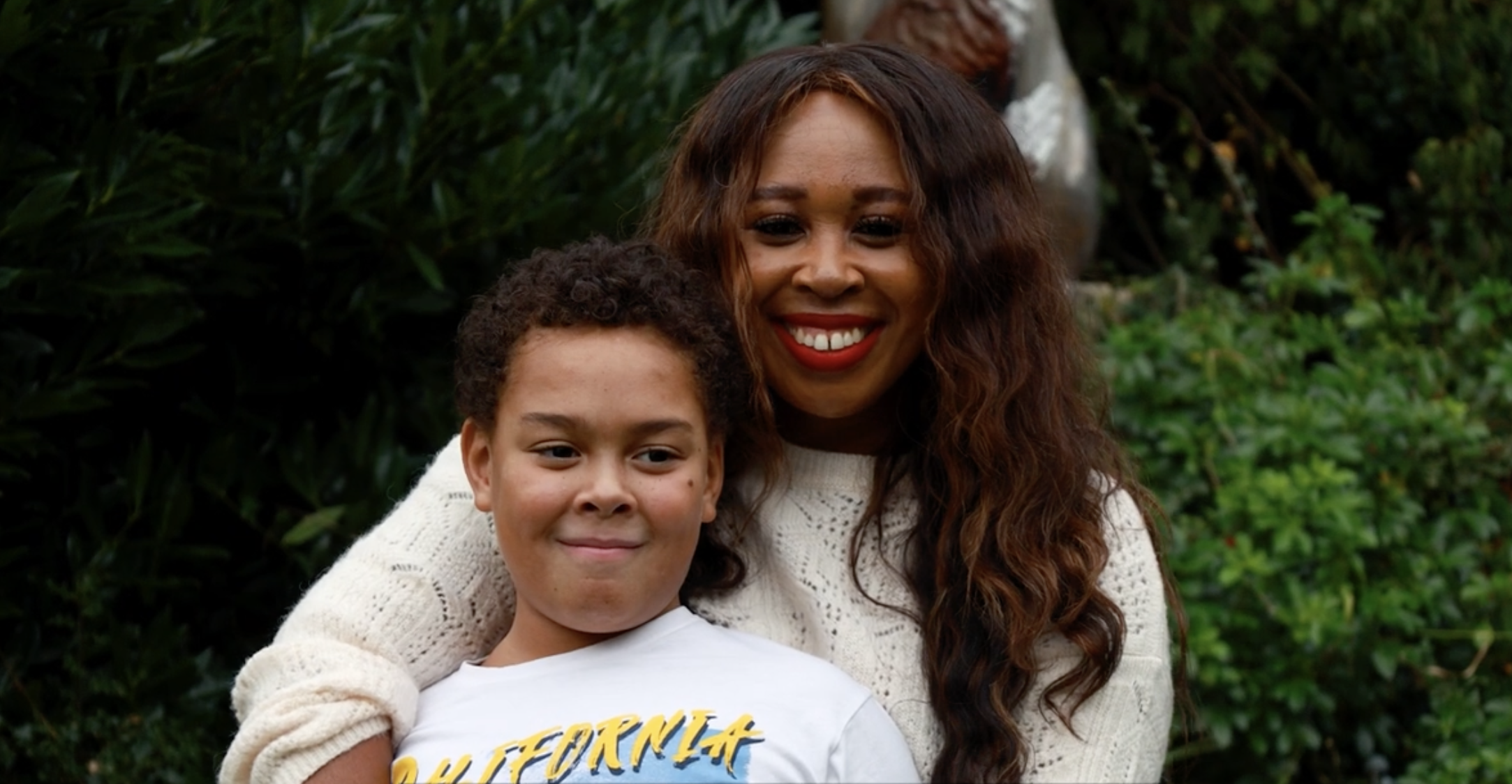 A woman with long wavy hair and bright red lipstick is smiling while hugging a young boy with short curly hair, both are outdoors with greenery in the background.
