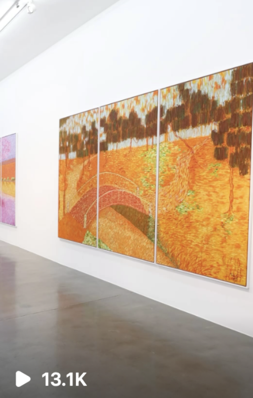 Three-panel painting of an orange autumn landscape with trees and a winding path, displayed in a gallery.