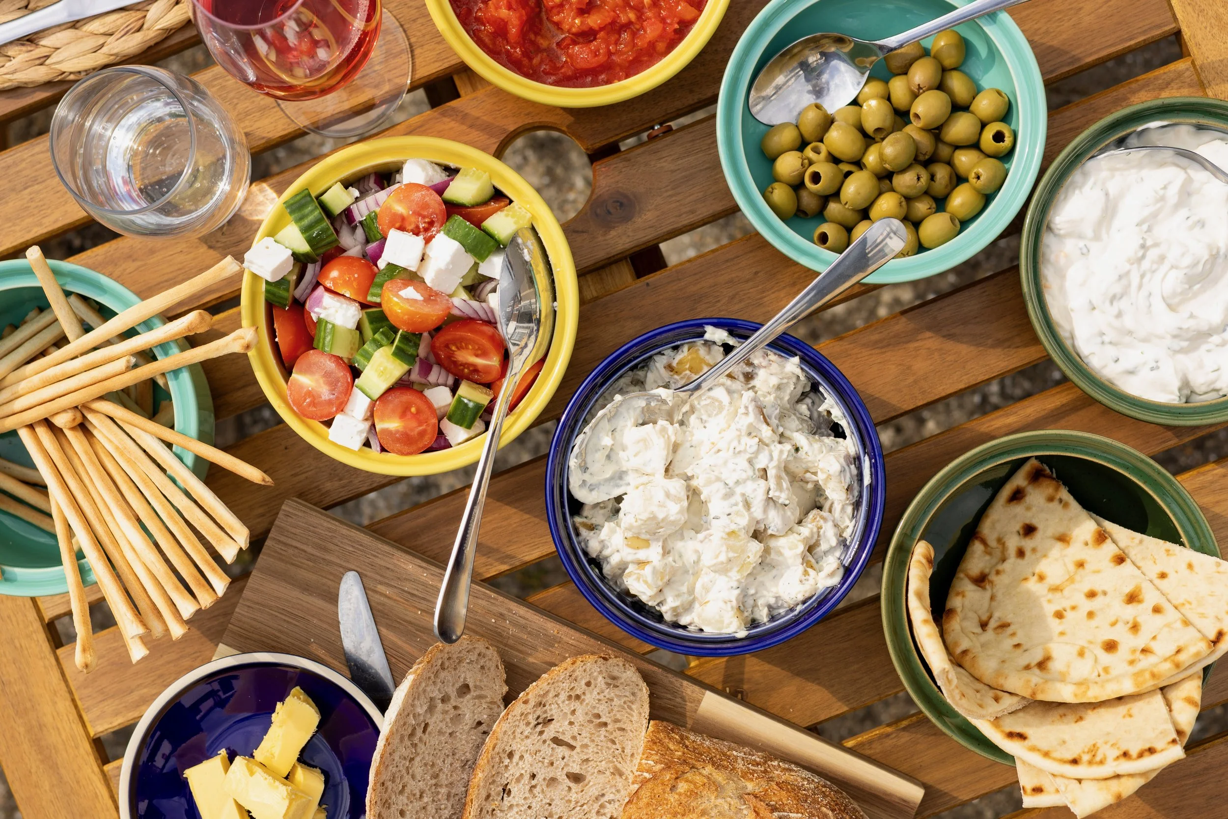 Assorted appetizers and dips on a wooden table, including a salad with cherry tomatoes, cucumbers, red onion, and feta cheese, bowls of green olives, white creamy dip, bread slices, and breadsticks, with a glass of rosé wine.