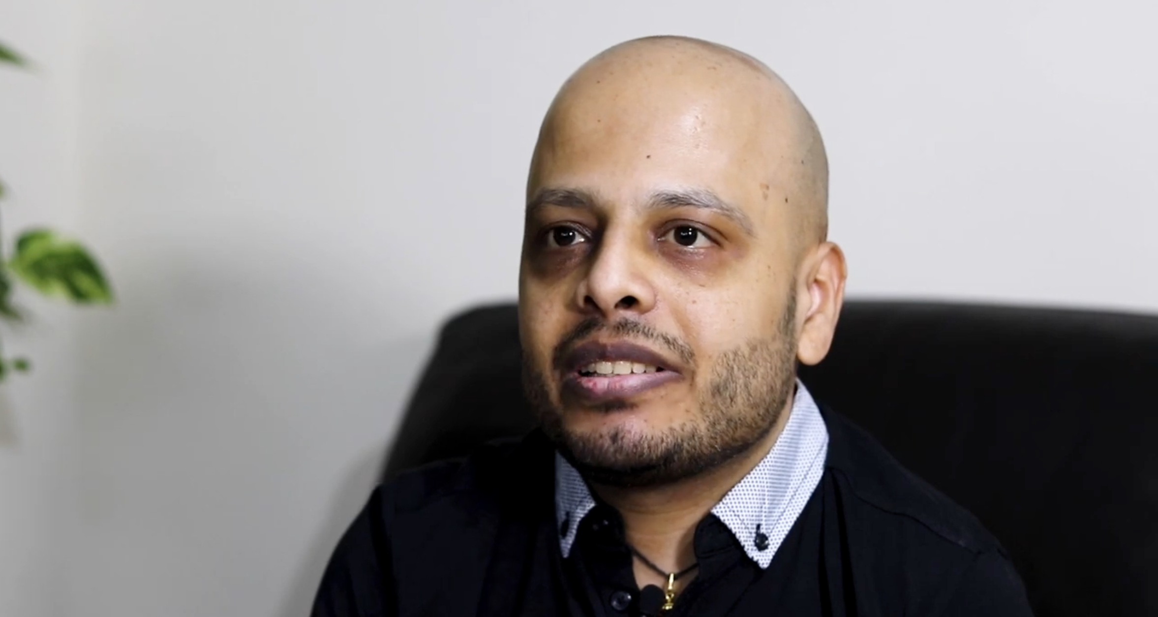 A close-up of a man with a shaved head and facial hair, sitting in an office chair, wearing a dark blazer, a button-up shirt with a subtle pattern, and a necklace, in front of a plain white wall.