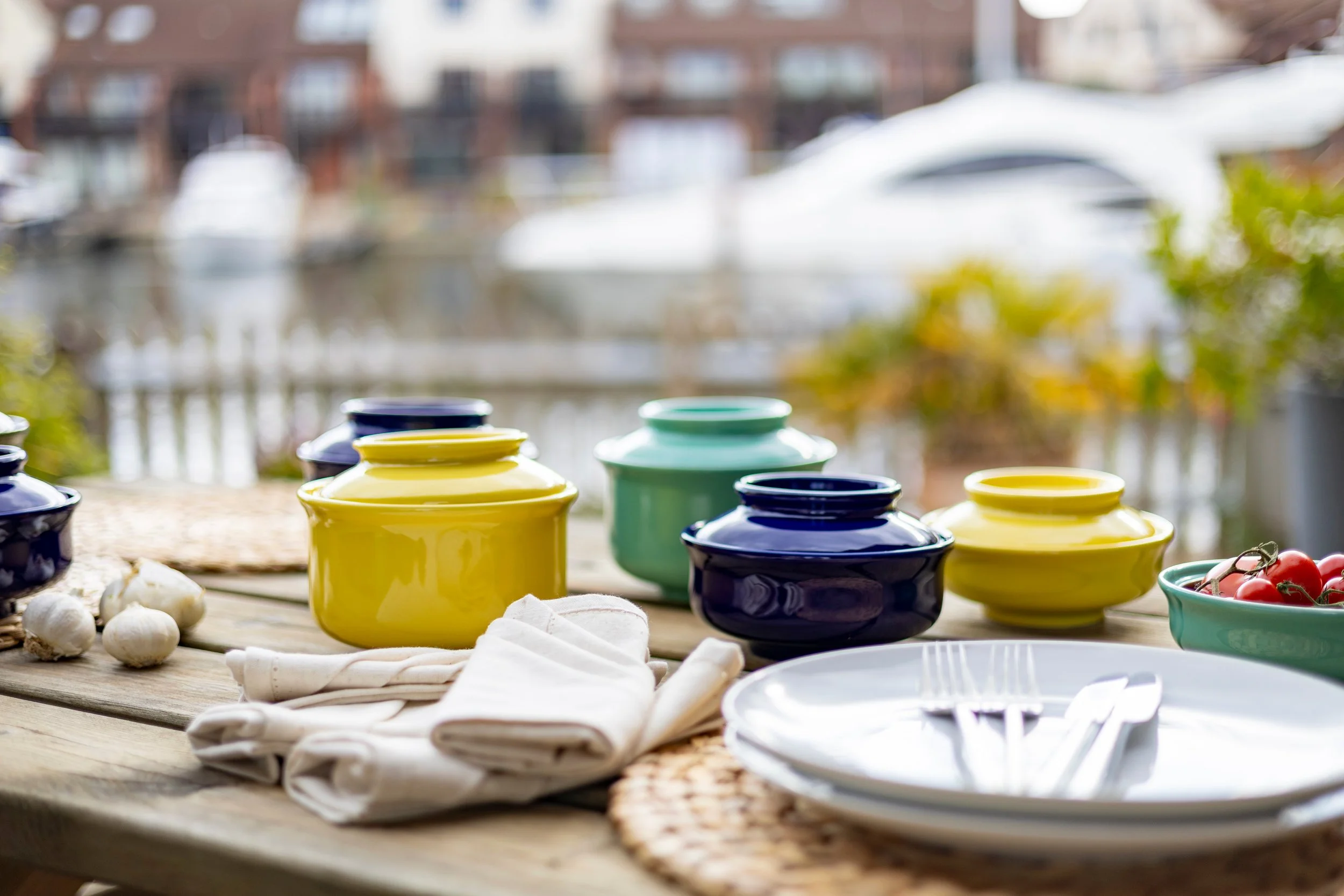 Colorful ceramic jars and bowls arranged on a wooden outdoor table, with garlic and tomatoes nearby, overlooking a marina with boats.