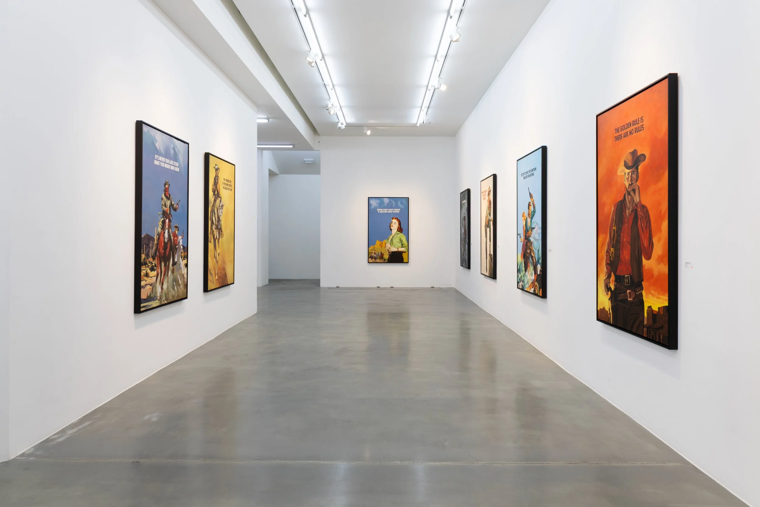 Gallery of framed colorful posters on white walls in an art gallery, with wooden flooring and ceiling lights.