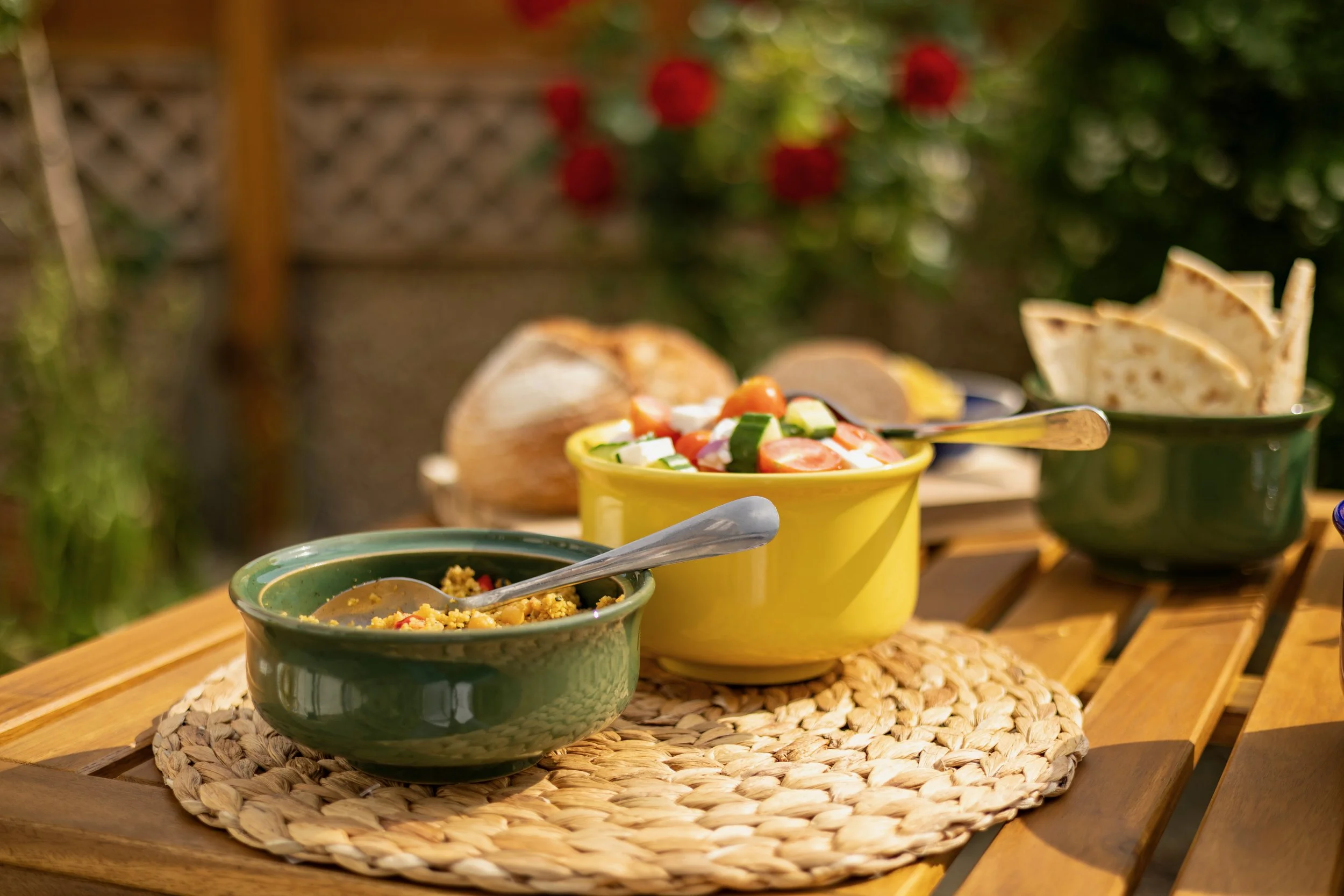 A table with bowls of colorful salad, hummus, and flatbread on a woven placemat outdoors with green plants and red flowers in the background.