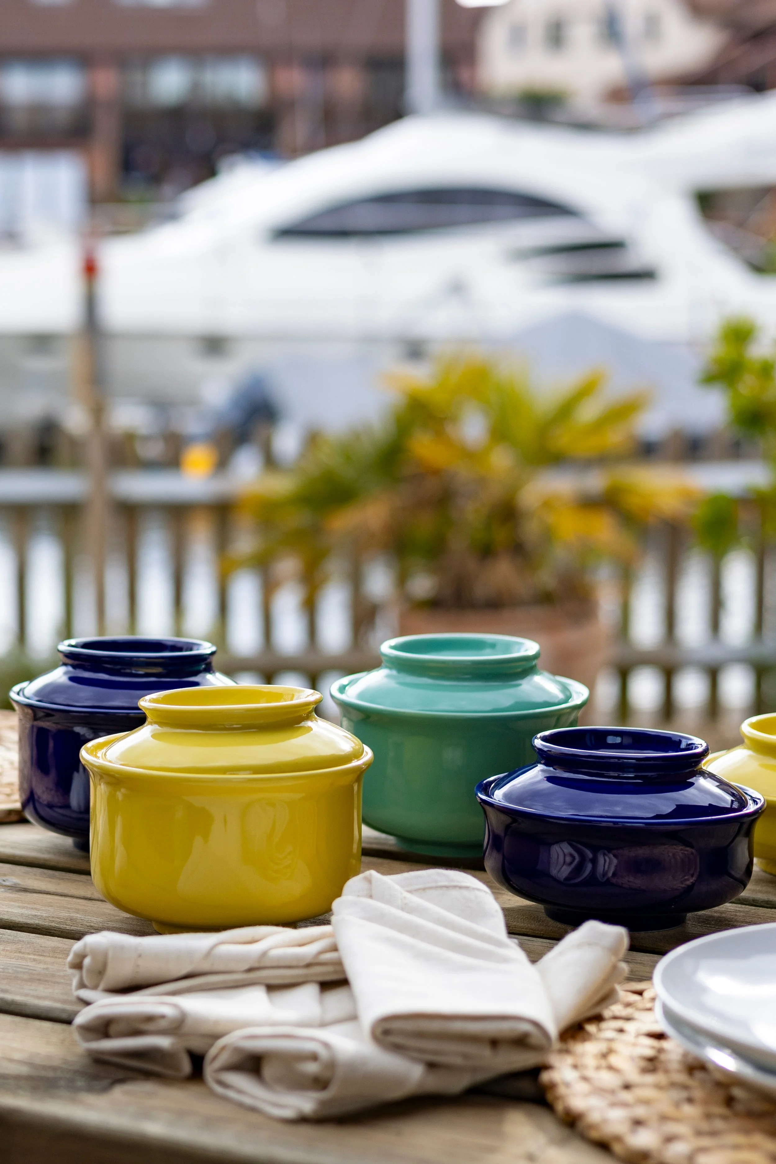 Colorful ceramic jars on a wooden table outdoors, with a blurred background including a boat, plants, and a deck.